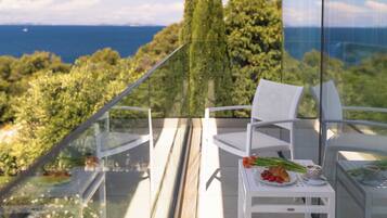 A balcony with a glass railing, white chairs, a small table with a plate of food, and a view of the sea and trees.