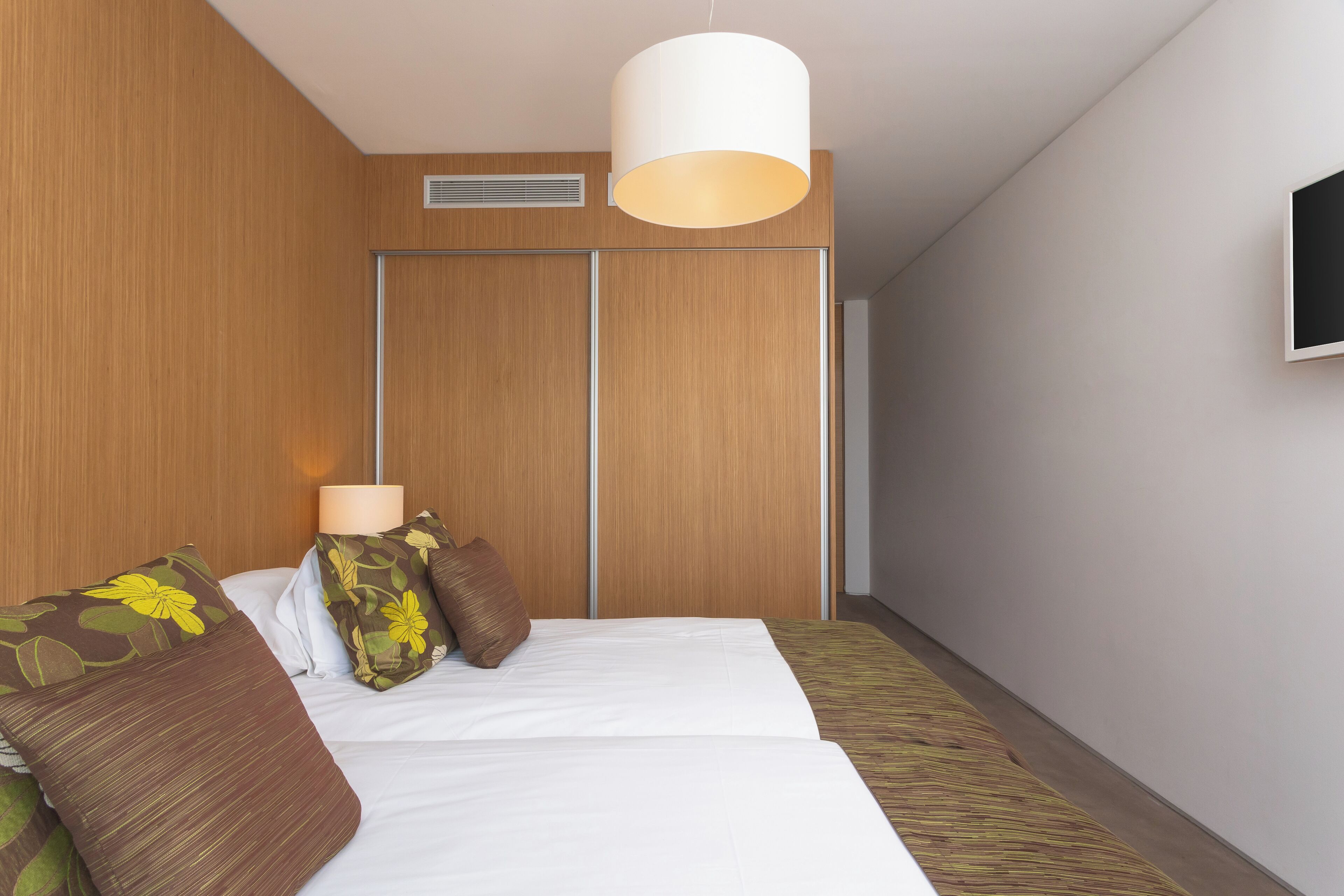 Una habitación de hotel con cama, almohadas, una lámpara de noche, armario y televisión en la pared.