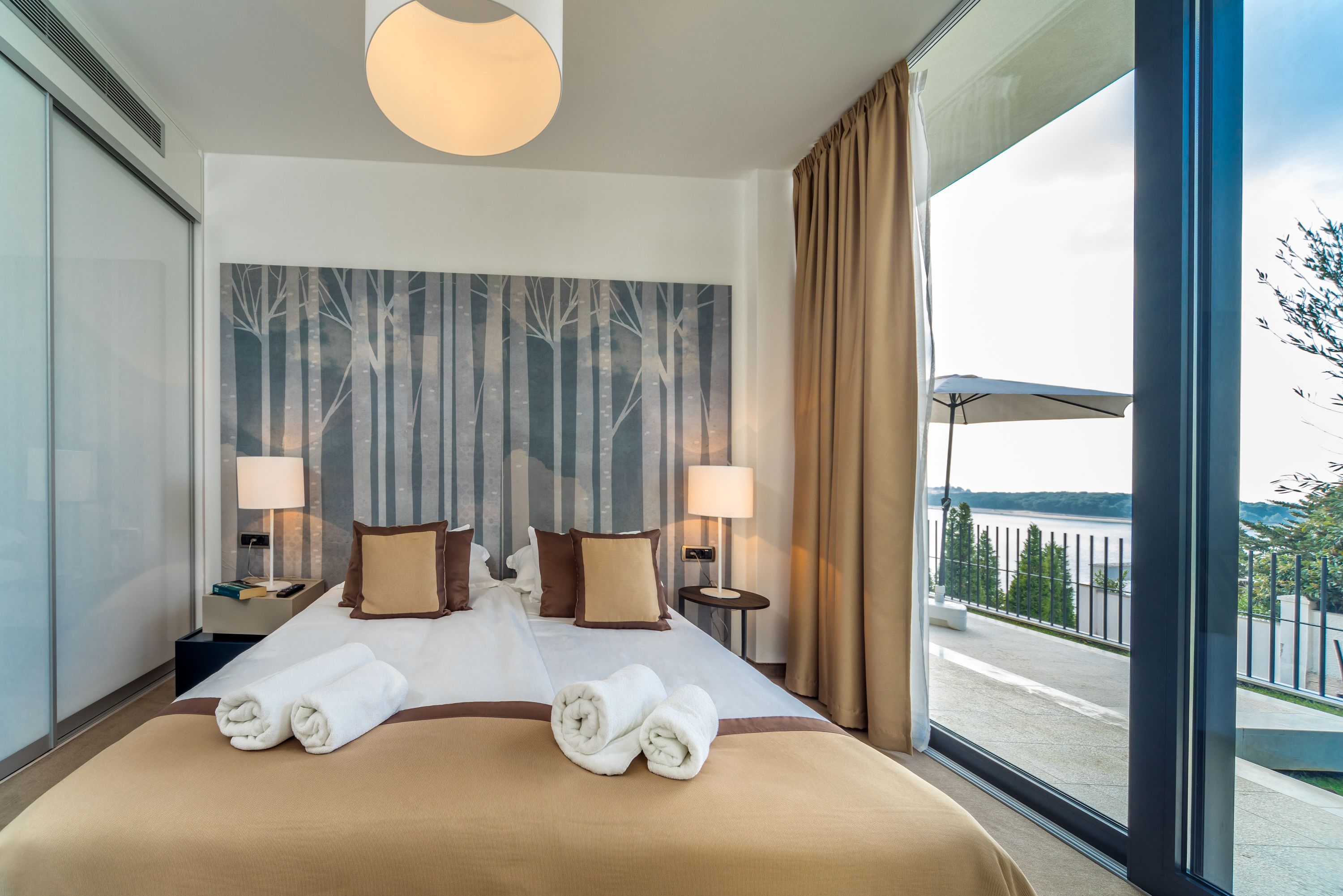 A hotel room with a large bed, two bedside tables with lamps, a view of a body of water through a window, and a cityscape in the distance.