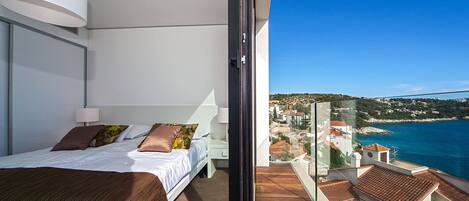 A bedroom with a bed, pillows, and a nightstand. There is a balcony with a view of the sea and buildings.