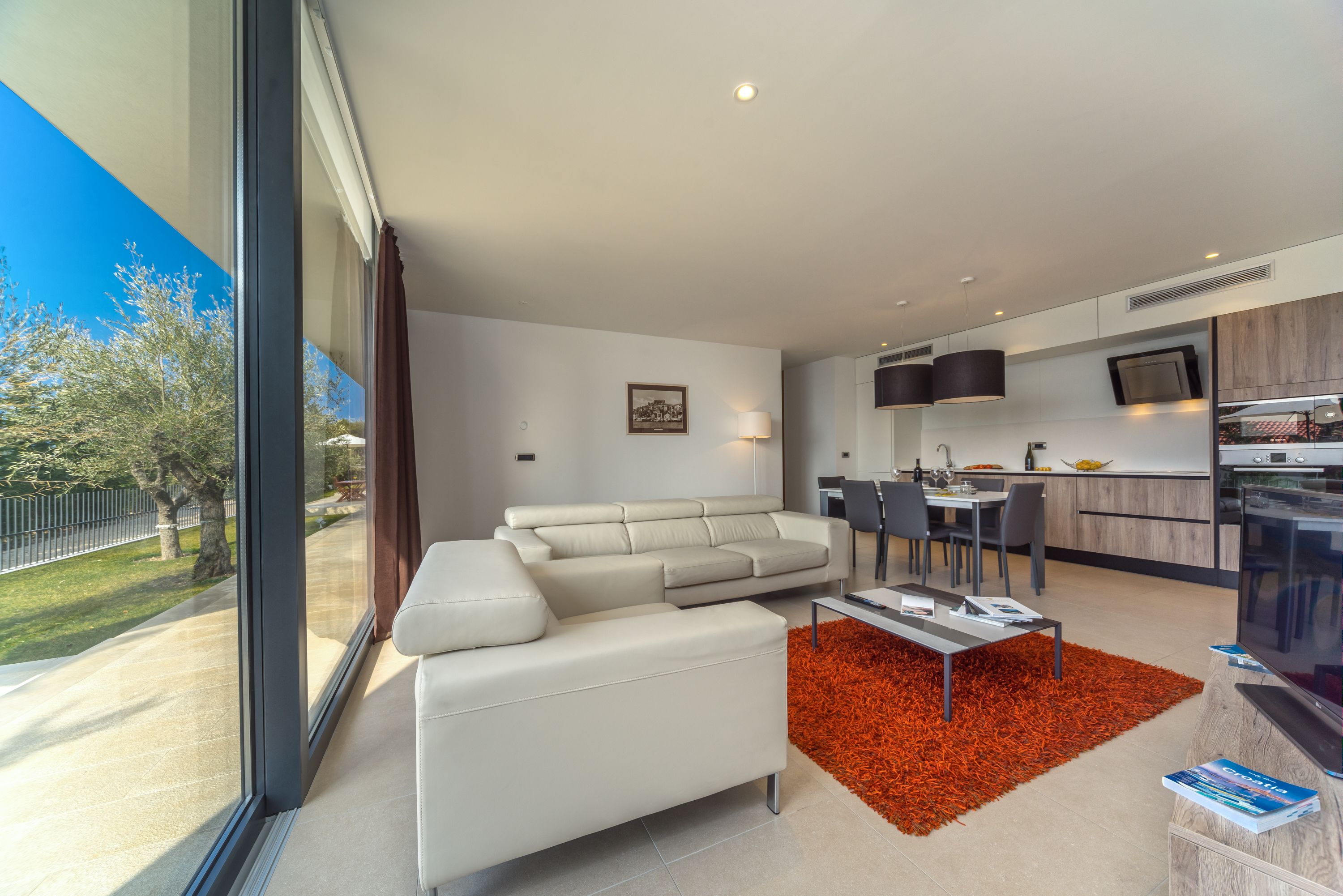 A modern living room with a large window, a sofa, a dining area, and a kitchen.