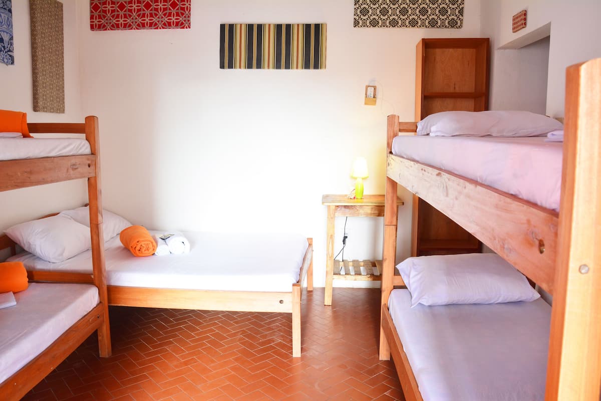 A dormitory room with bunk beds, a small table with a lamp, and a wall-mounted shelf.