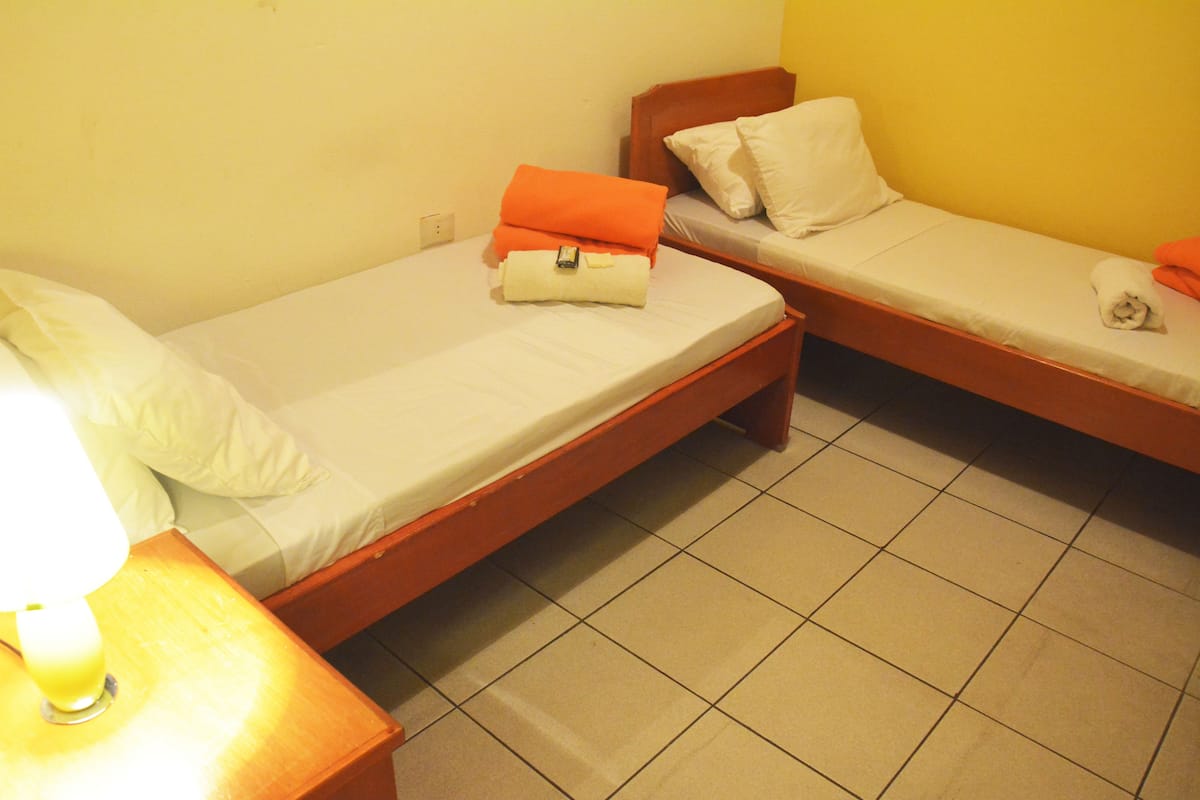 Two single beds with wooden frames and white bedding, a nightstand with a lamp, and a tiled floor.