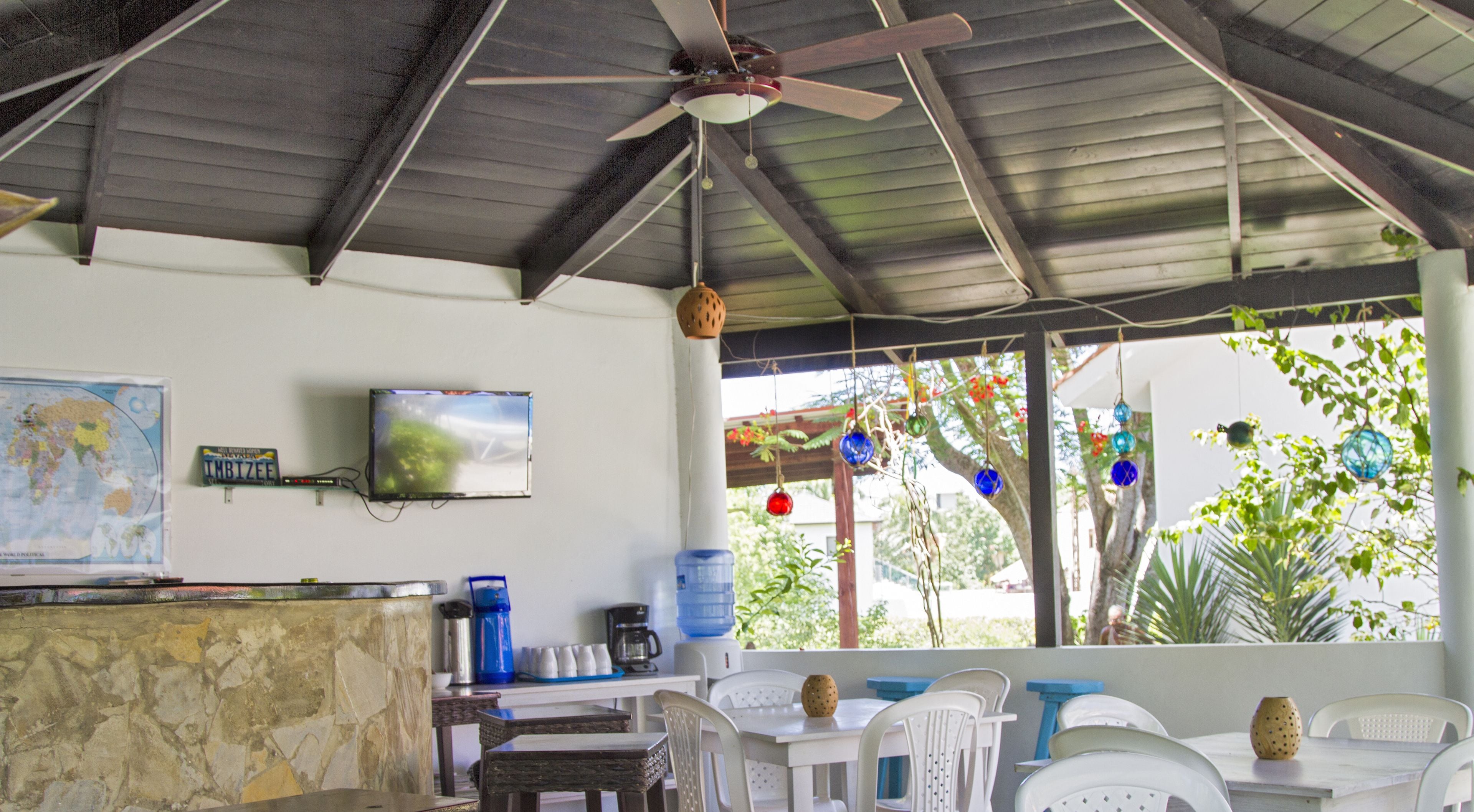 A covered outdoor seating area with a ceiling fan, a television mounted on the wall, and a view of greenery outside.