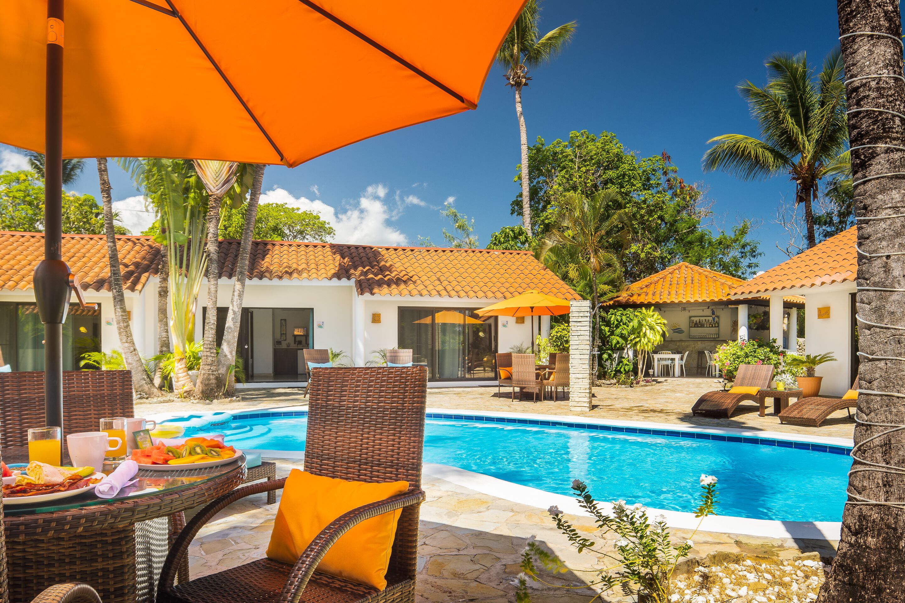 A poolside area with wicker chairs and orange umbrellas, a swimming pool, and palm trees.