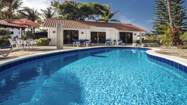 A swimming pool with clear blue water, surrounded by lounge chairs and a building with a tiled roof.