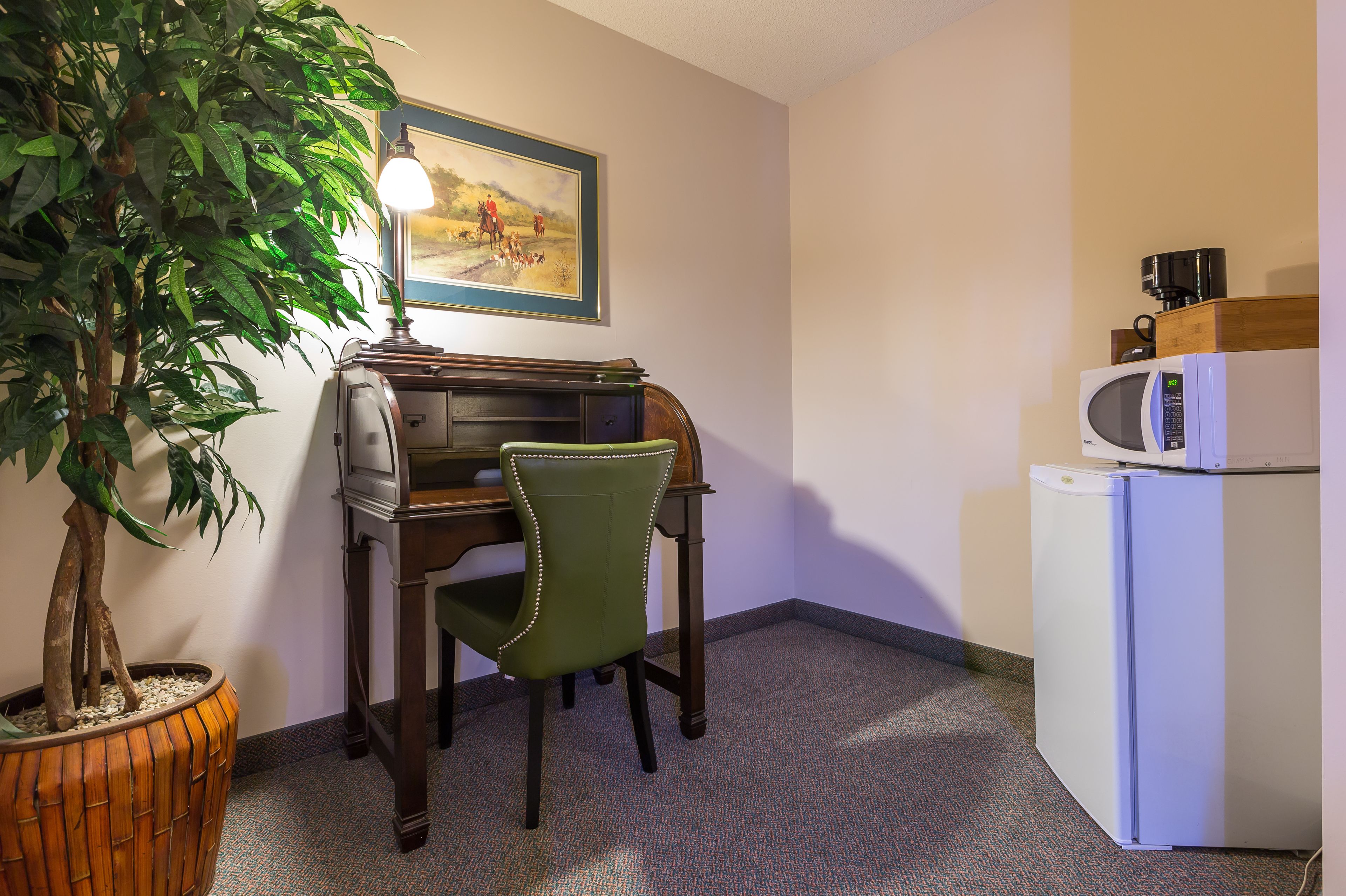 Une petite pièce comprenant un bureau, une chaise verte, une plante en pot, un four à micro-ondes et un réfrigérateur.