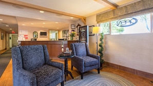 A lobby with two patterned armchairs, a reception desk, a clock, and a window with a view of trees. - Grama's Inn (Prince George)