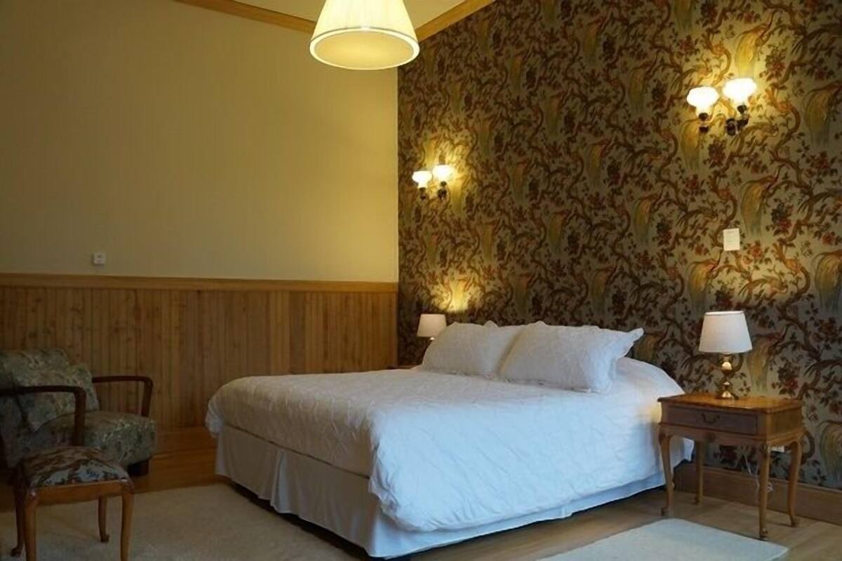 Un dormitorio con una cama grande, dos lámparas de pared, una silla y una mesita de noche con lámpara.