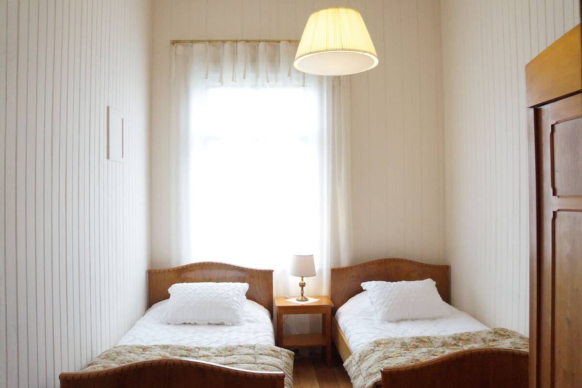 Una habitación con dos camas de madera, una mesita de noche con lámpara y una ventana con cortinas.