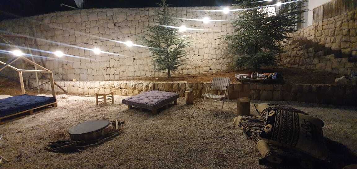 Un patio al aire libre con pared de piedra, luces colgantes, banco de madera, zona de estar acolchada y una mesita.