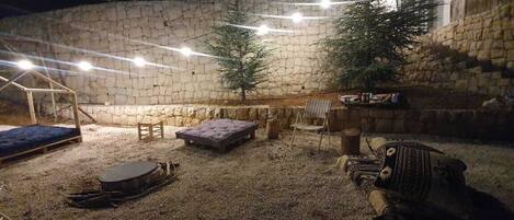 Un patio al aire libre con pared de piedra, luces colgantes, banco de madera, zona de estar acolchada y una mesita.