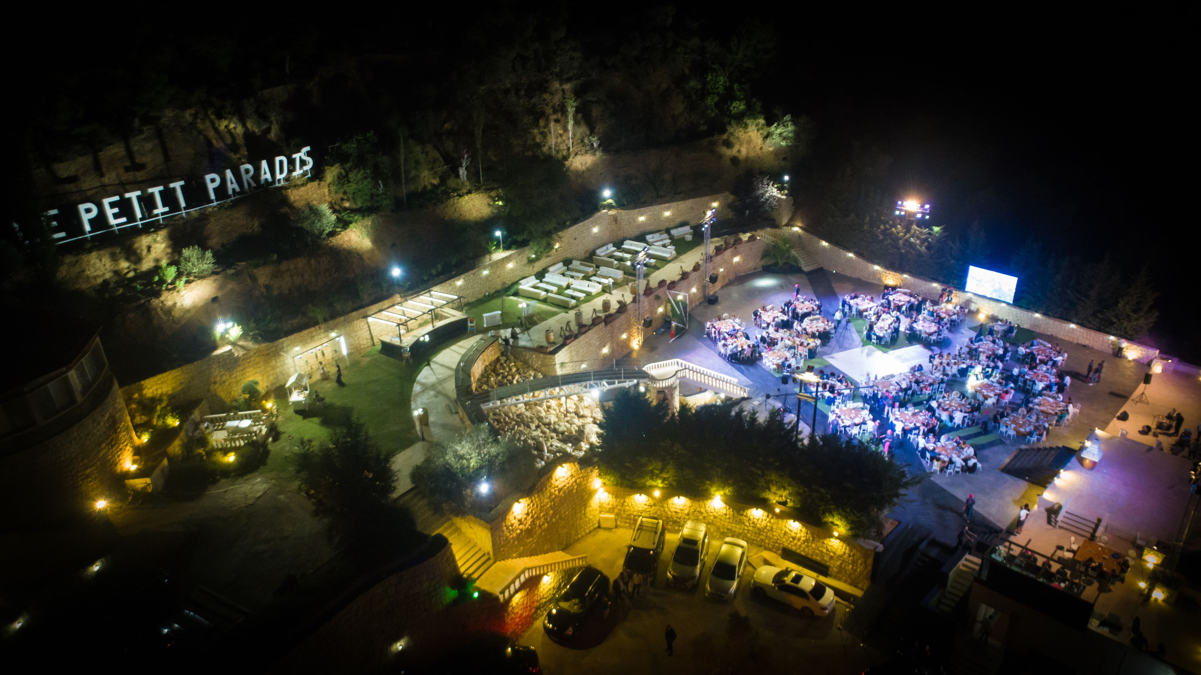Vista aérea de un espacio exterior iluminado para eventos, con varias mesas y sillas, rodeado de muros de piedra y senderos.