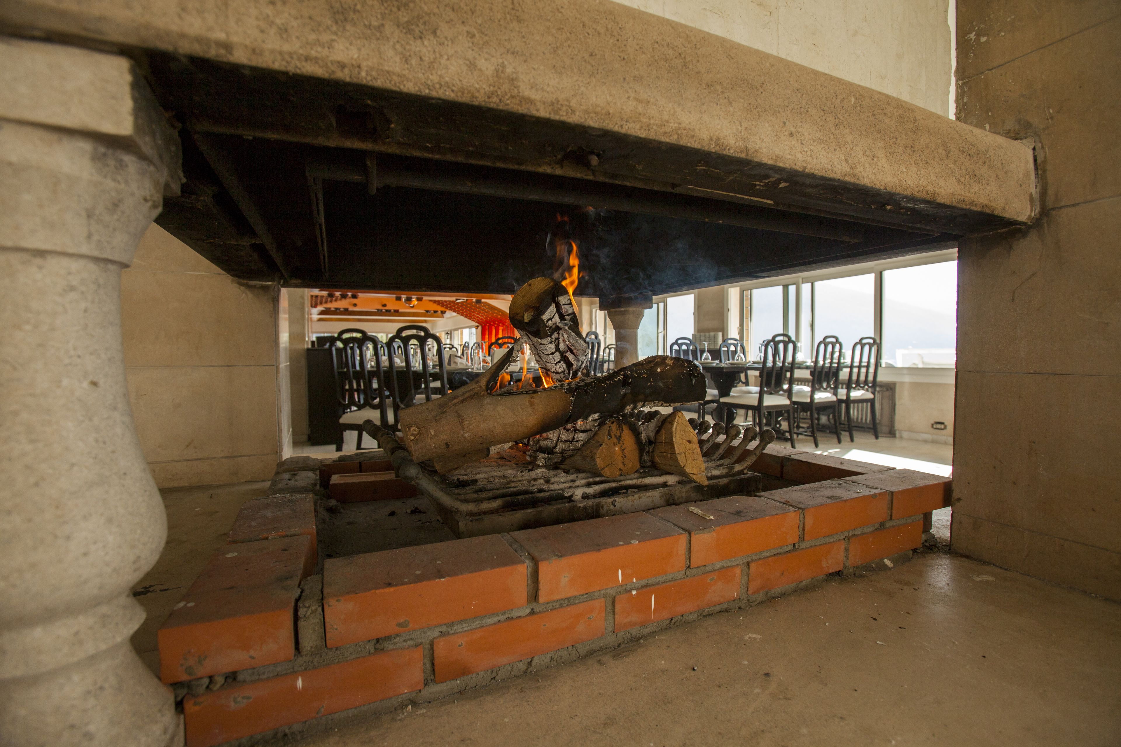 A brick fireplace with burning logs, located in a room with a concrete pillar and a view of a dining area with chairs and tables.