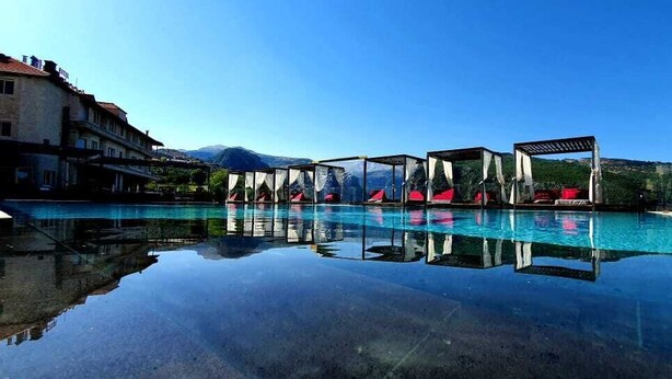 A swimming pool with clear water, surrounded by buildings and a mountain in the background. - Le Tournant Hotel & Resort (Ehden)