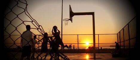 A basketball court at sunset with a chain-link fence and a basketball hoop.
