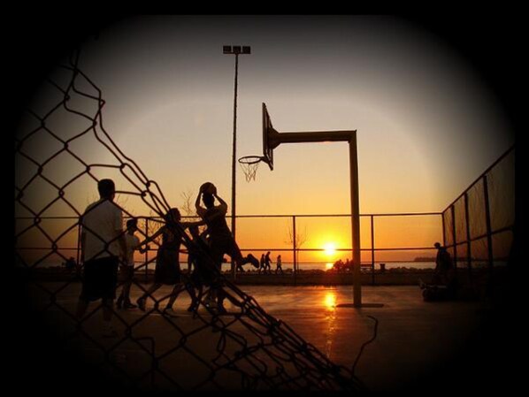 Una cancha de baloncesto al atardecer, con una cerca de malla y un aro de baloncesto.