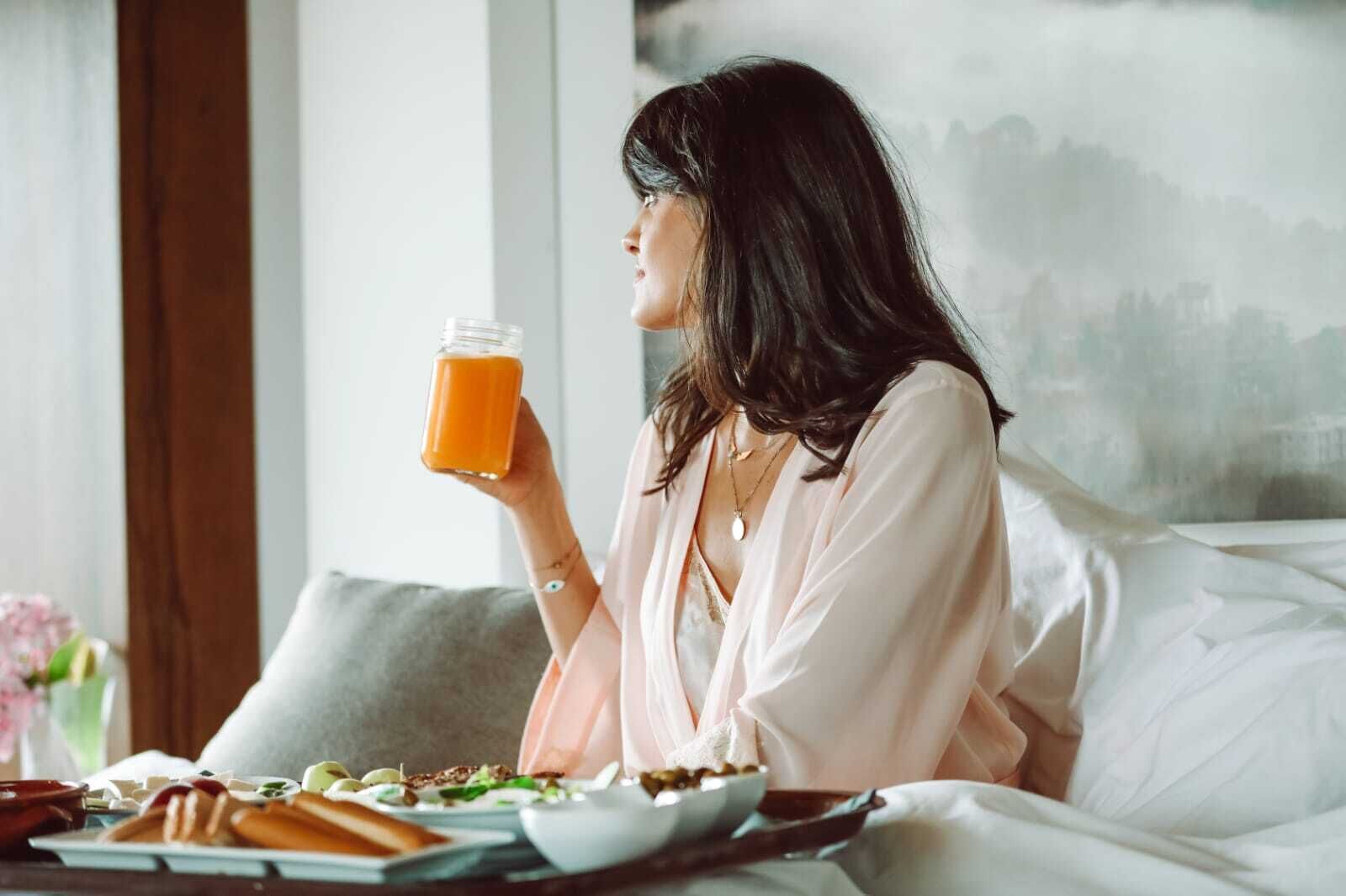Une personne assise sur un lit, avec un plateau de nourriture et un verre de jus d’orange.