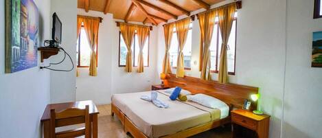 A bedroom with a wooden bed, a desk, and a chair. There are curtains on the windows, and a painting is on the wall.