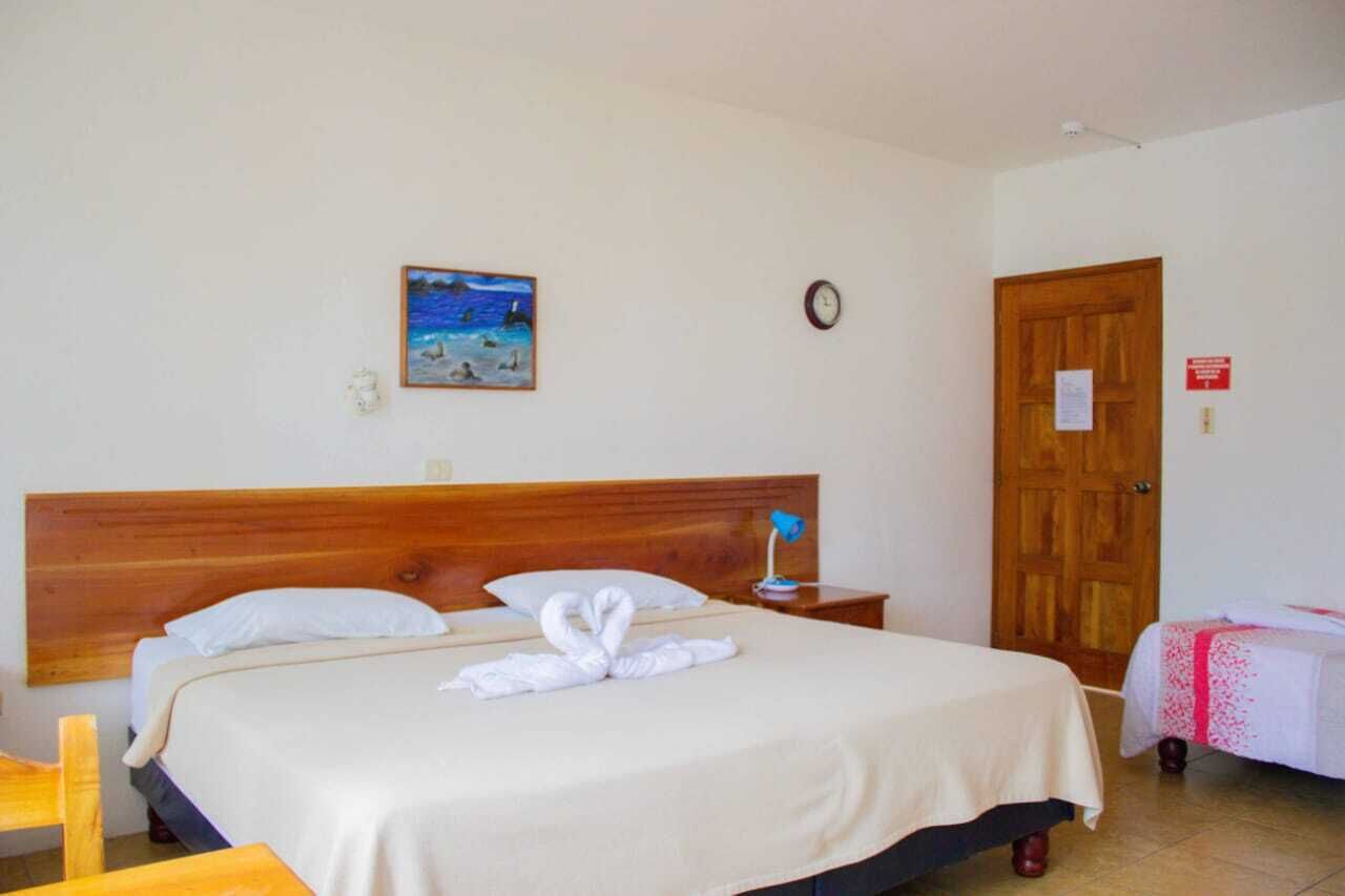 Quarto de hotel com cabeceira de madeira, cama com lençóis brancos, pintura de uma cena de praia, relógio e porta de madeira.