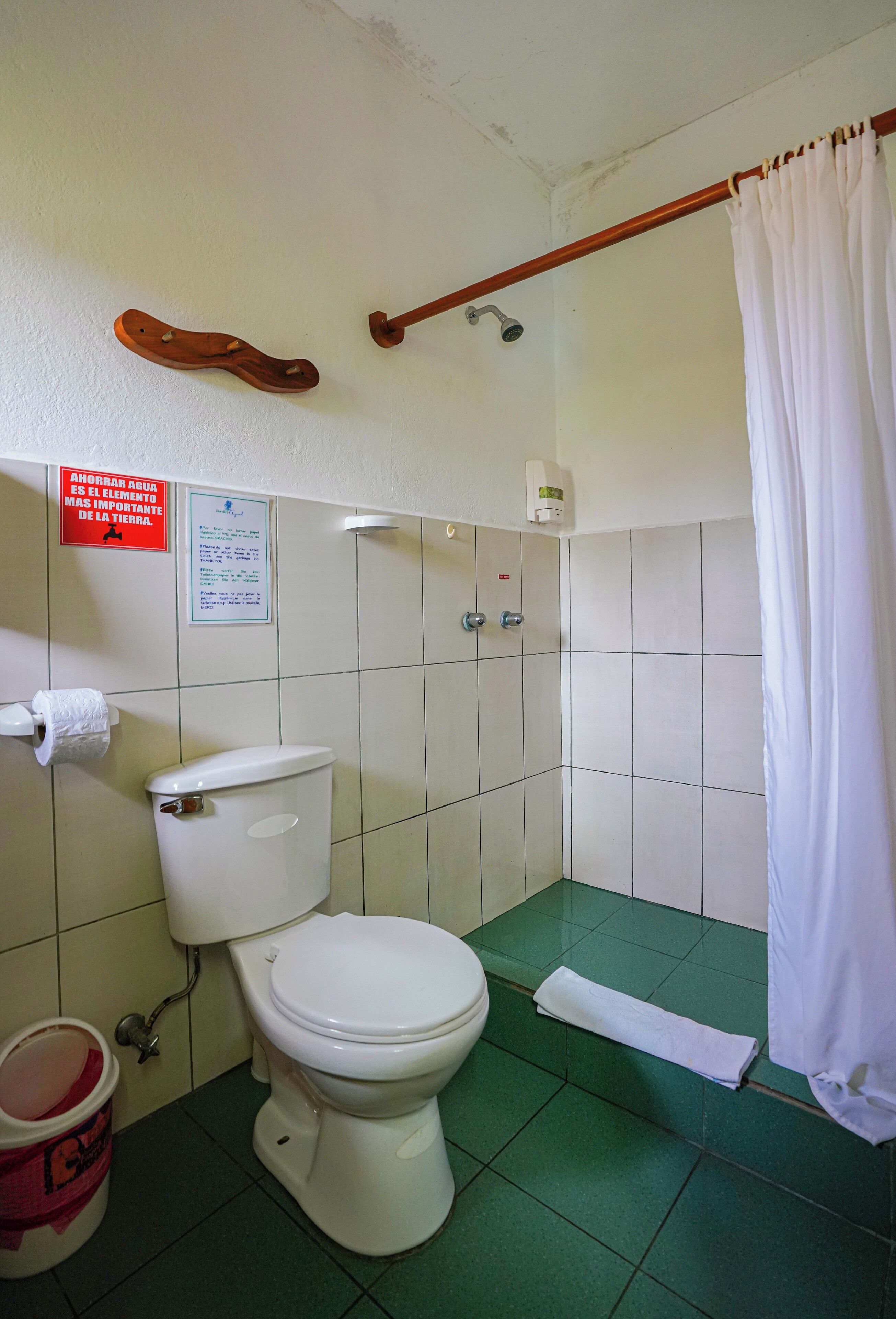 A small bathroom with a toilet, a shower, and a curtain. There is a sign on the wall and a bucket on the floor.