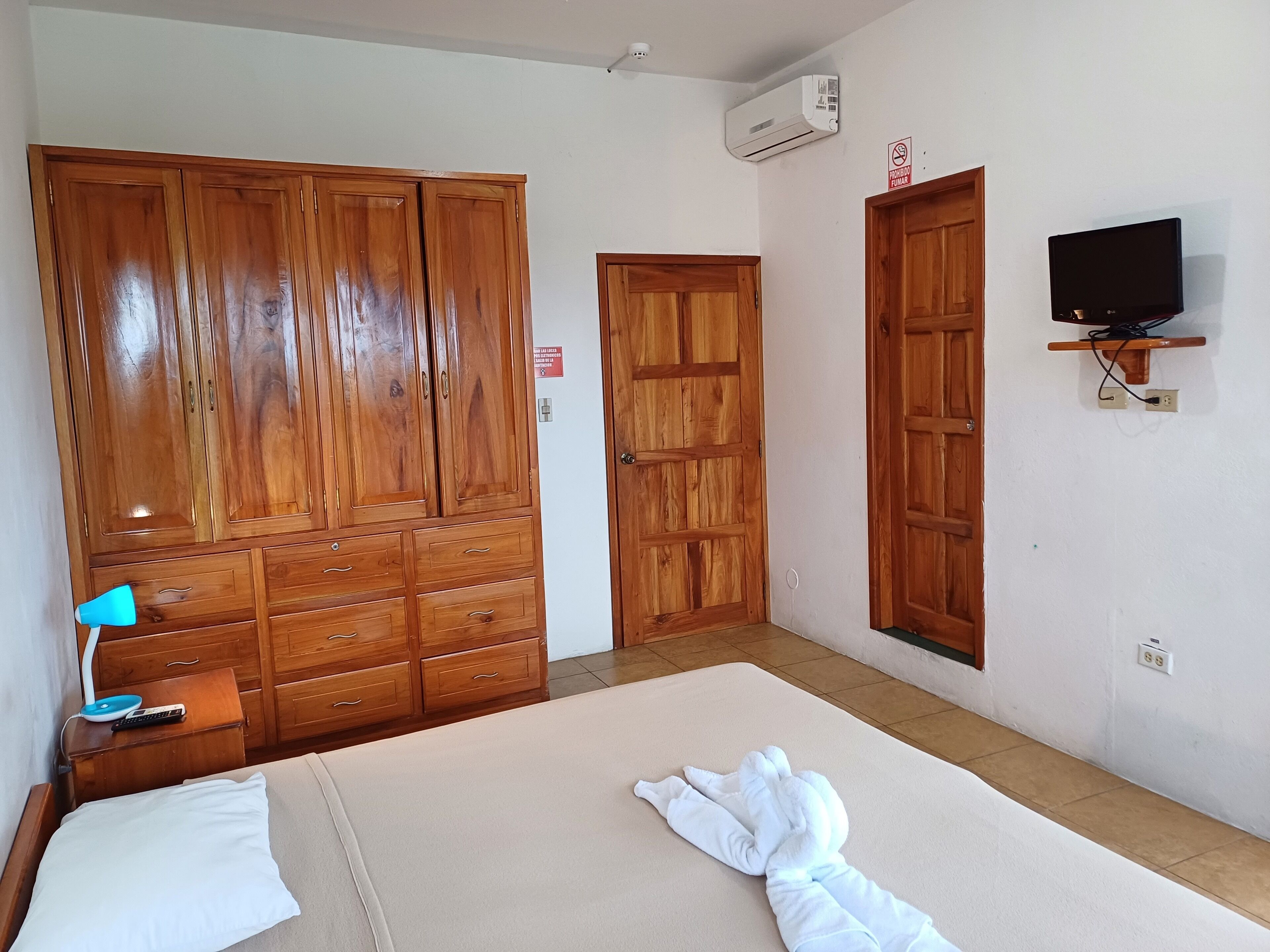 A bedroom with a large wooden wardrobe, a bed with white bedding, a blue lamp, and a television mounted on the wall.