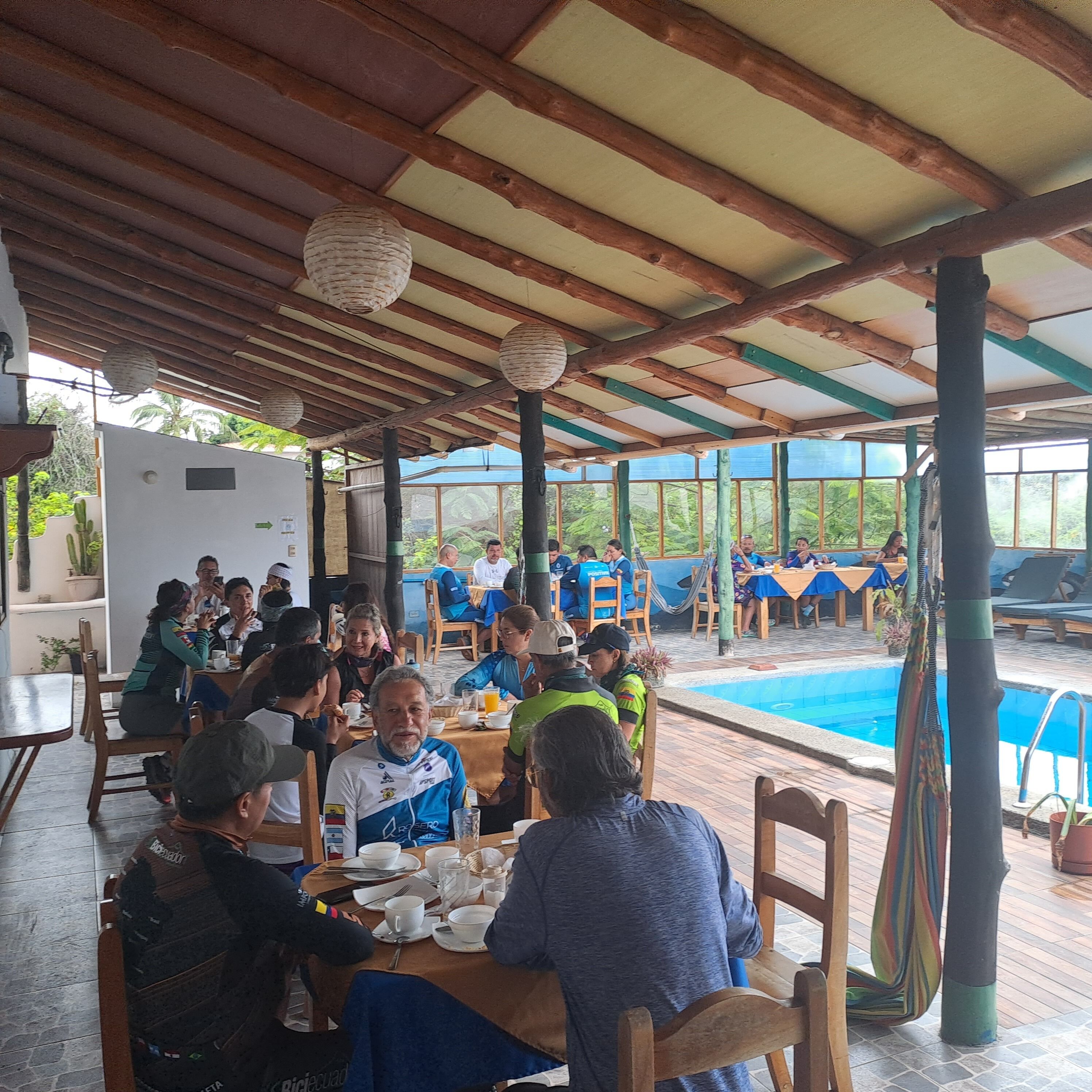 Un espace extérieur abrité, aménagé pour les repas, avec des tables et des chaises en bois, entouré d’une piscine et de verdure.