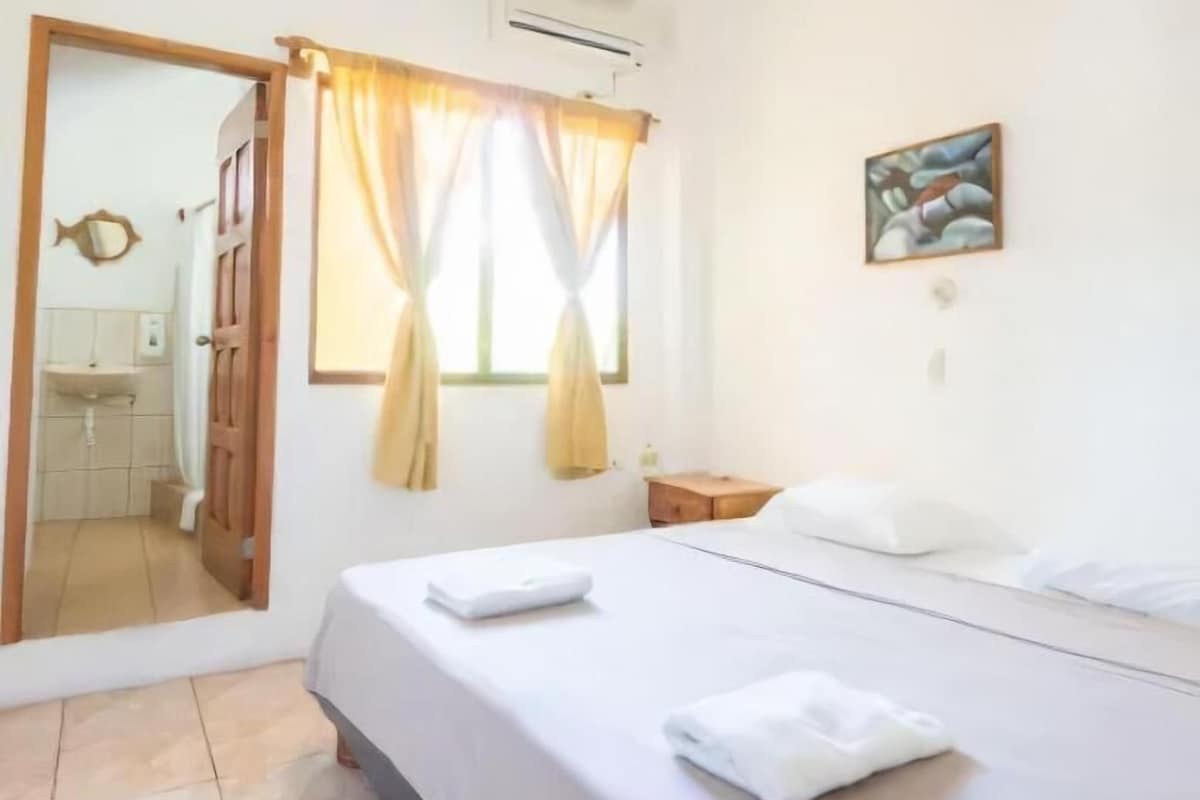 A hotel room with a bed, a nightstand, a painting, and a bathroom visible through an open door.