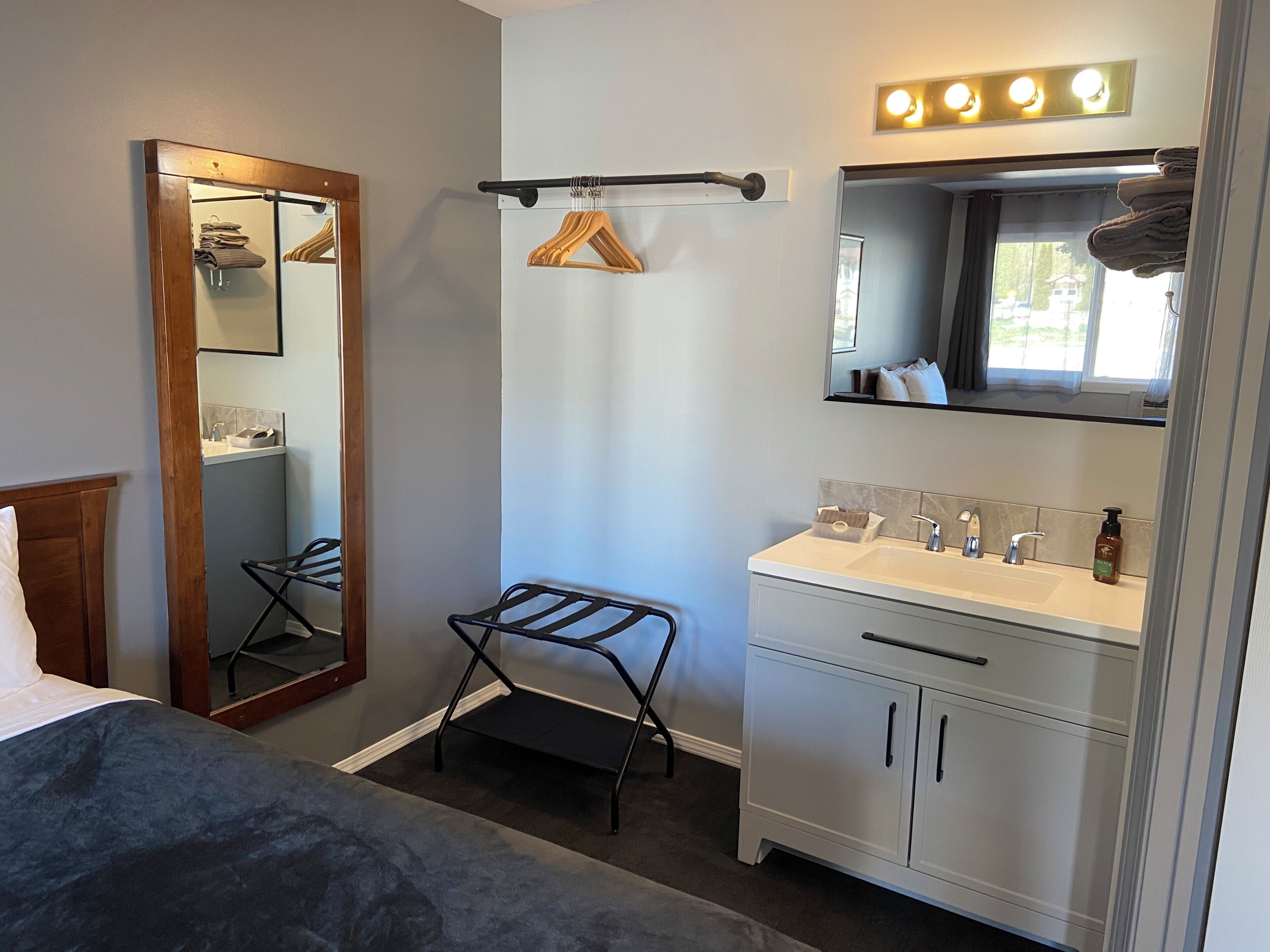 A hotel room with a bed, a bathroom with a sink and mirror, a hanging clothes rack, and a window with curtains.