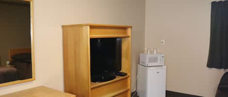 A hotel room with a wooden TV stand, a small white refrigerator, a microwave, a wooden desk, and a mirror.