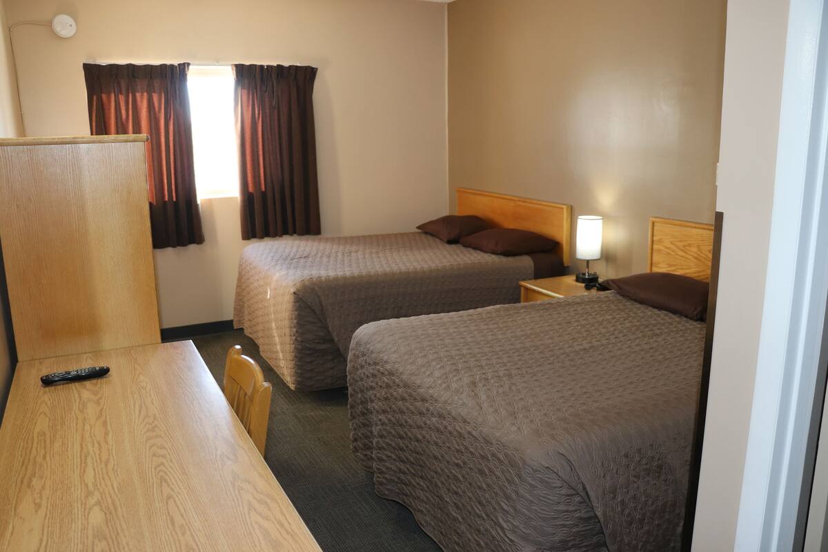 A hotel room with two beds, a wooden desk, a chair, a lamp, and a window with curtains.