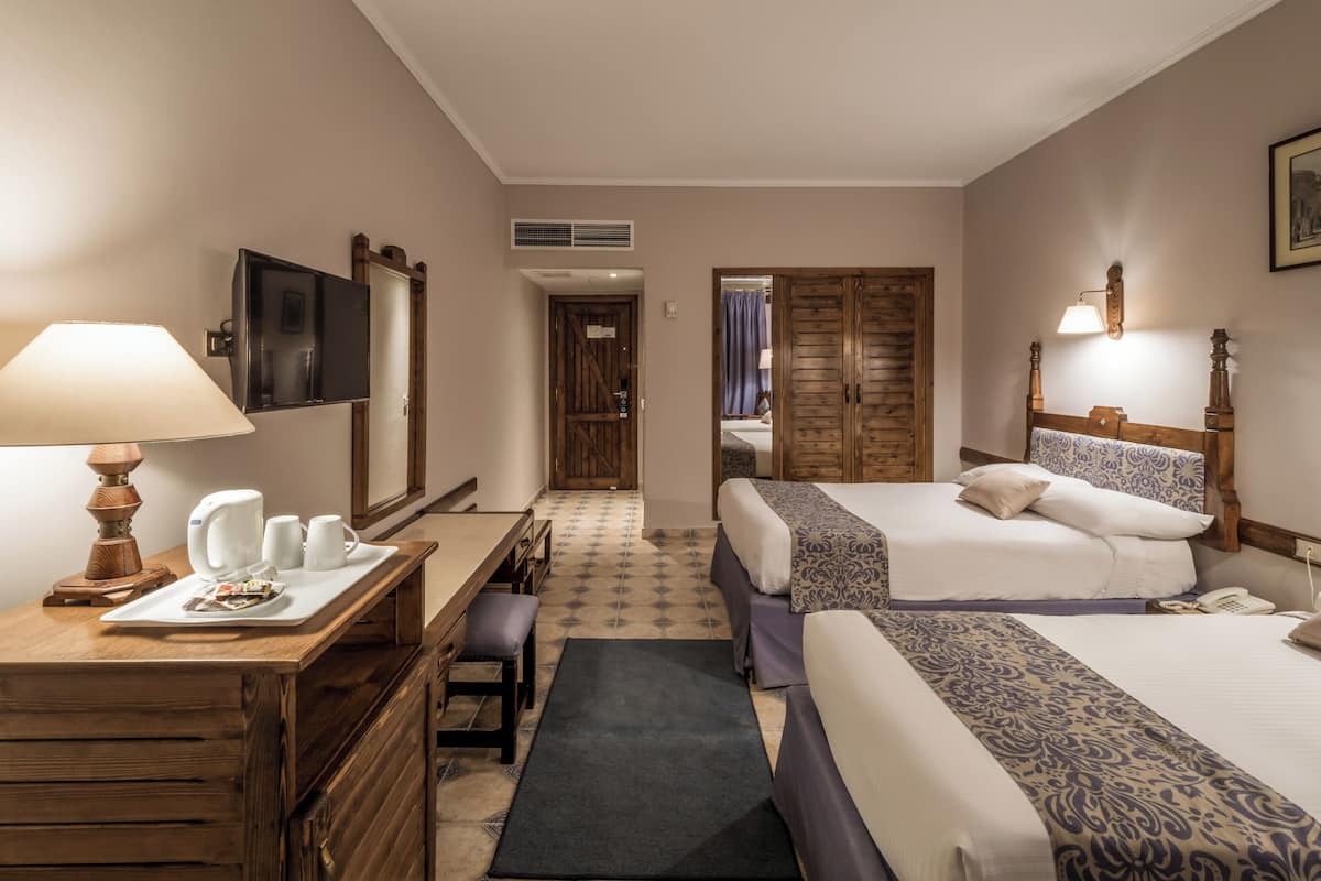 A hotel room with two beds, a desk with a lamp, a coffee maker, and a TV on the wall.