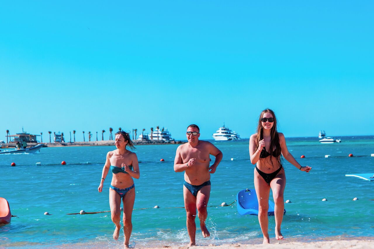 Tres personas corriendo en una playa, con embarcaciones y construcciones al fondo.