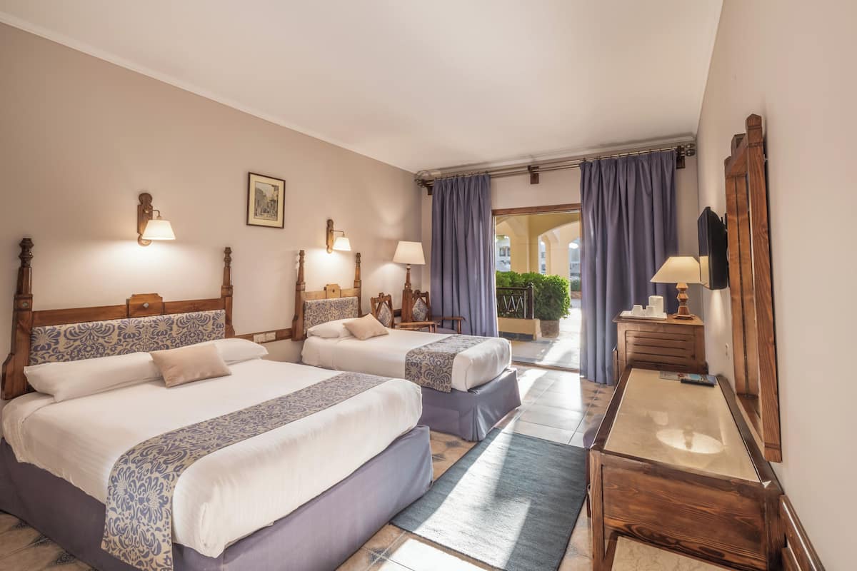 A hotel room with two beds, a wooden headboard, a nightstand, a mirror, and a view of the courtyard.
