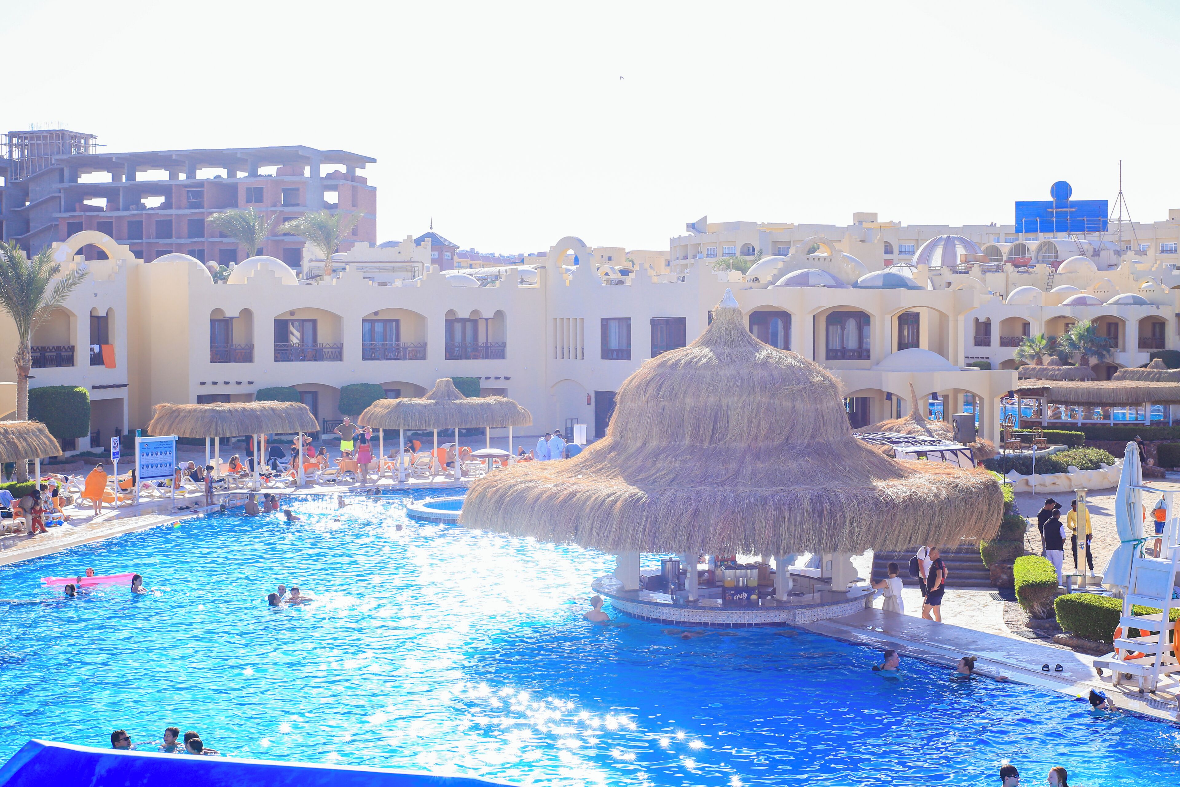 A swimming pool with a central island, surrounded by buildings with arched windows and a thatched-roof structure.