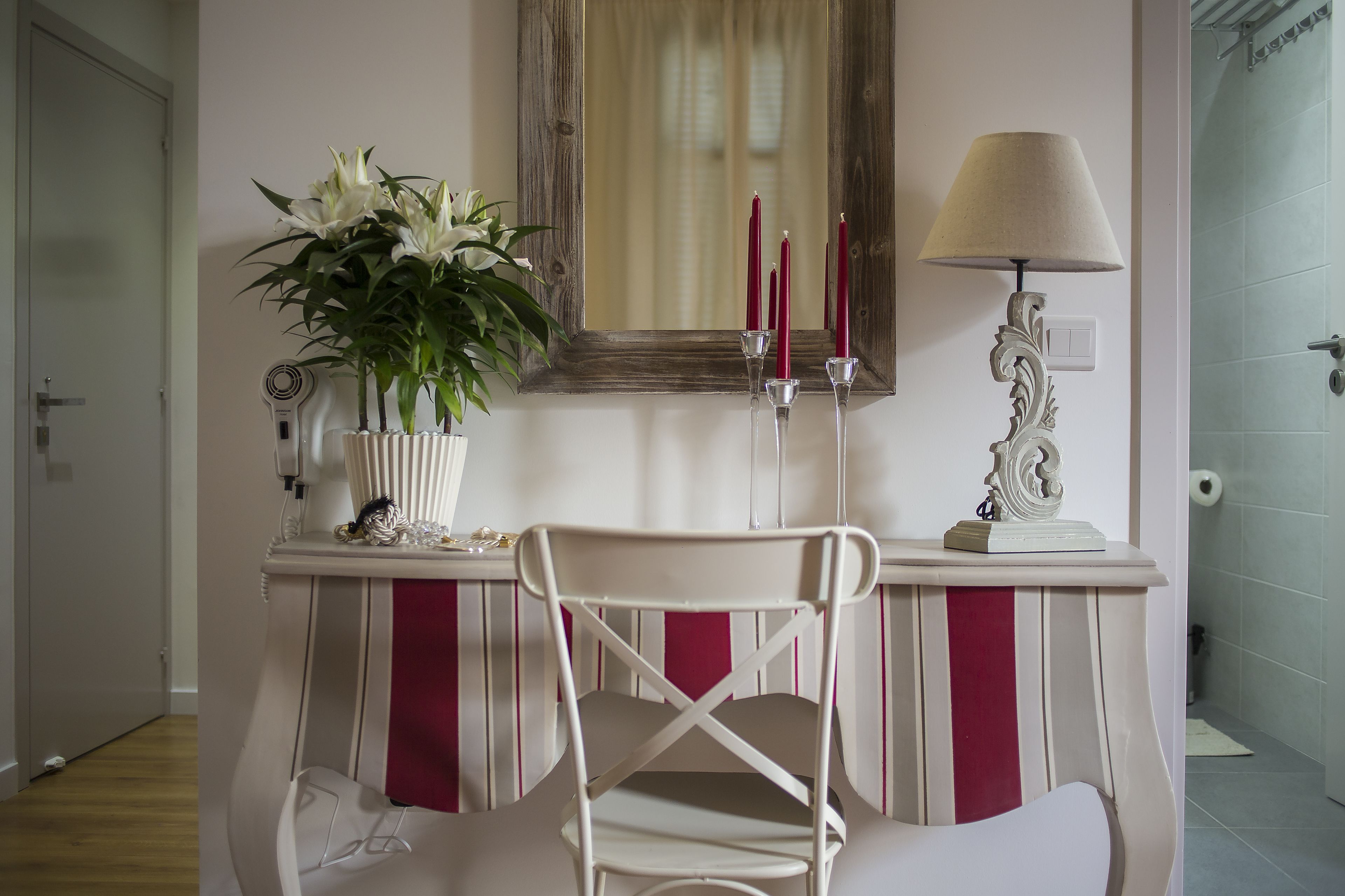 A room with a white chair, a mirror, a vase with white flowers, and a lamp.