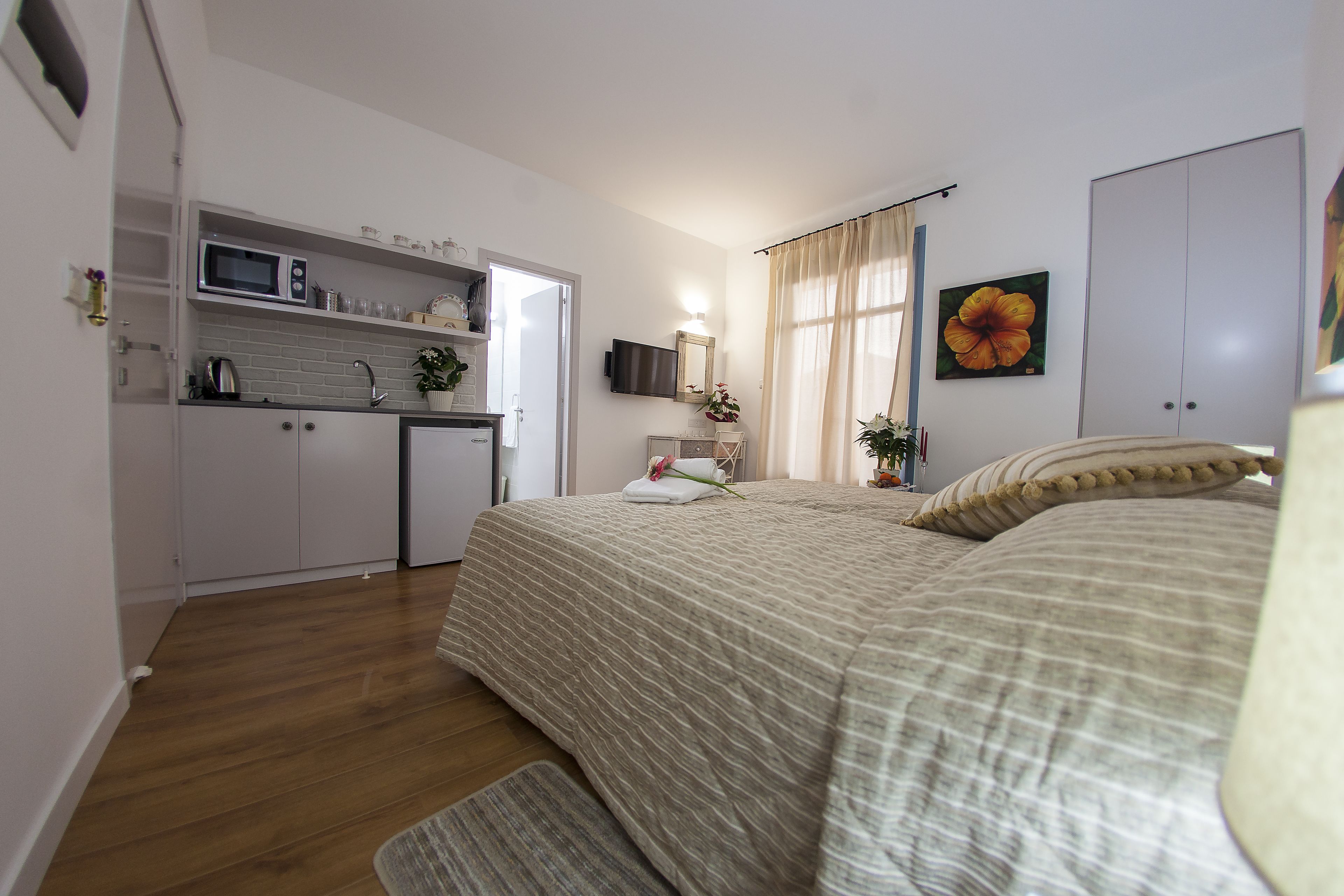 A compact bedroom with a bed, a bedside table, a small desk with a chair, a window with curtains, and a kitchenette with a microwave and sink.