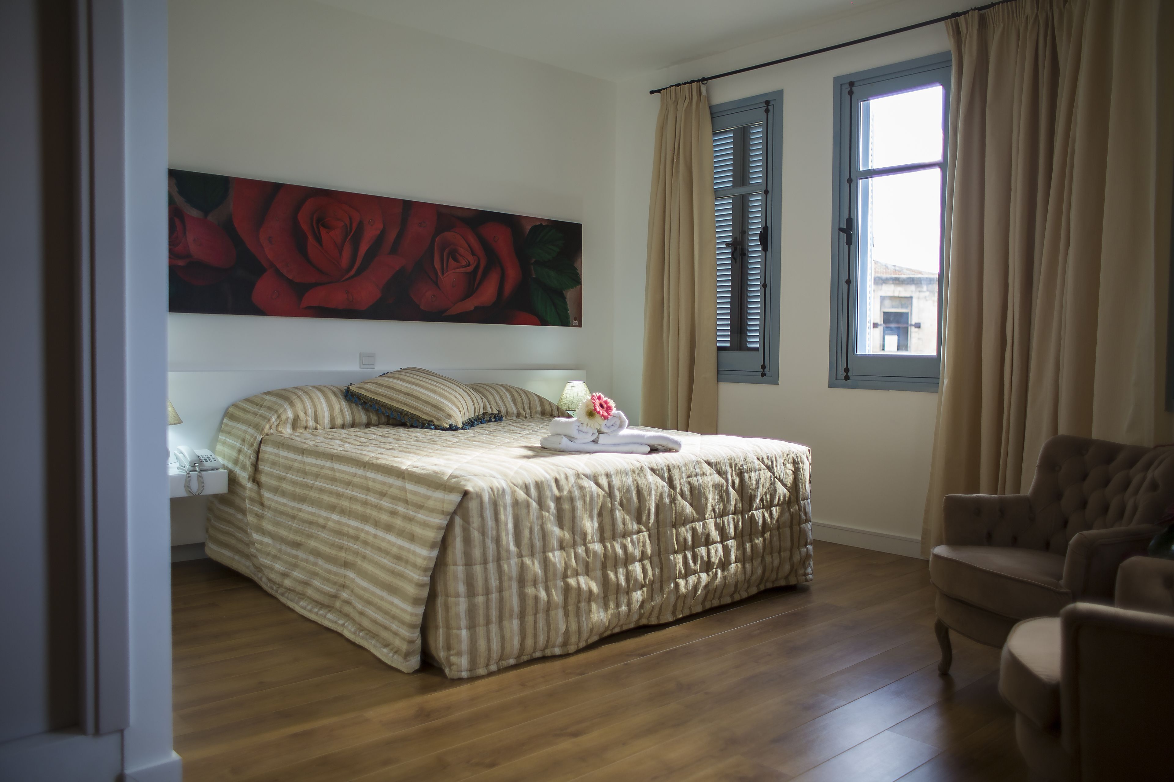 A bedroom with a large bed, a nightstand, a chair, a window with curtains, and a painting of roses on the wall.