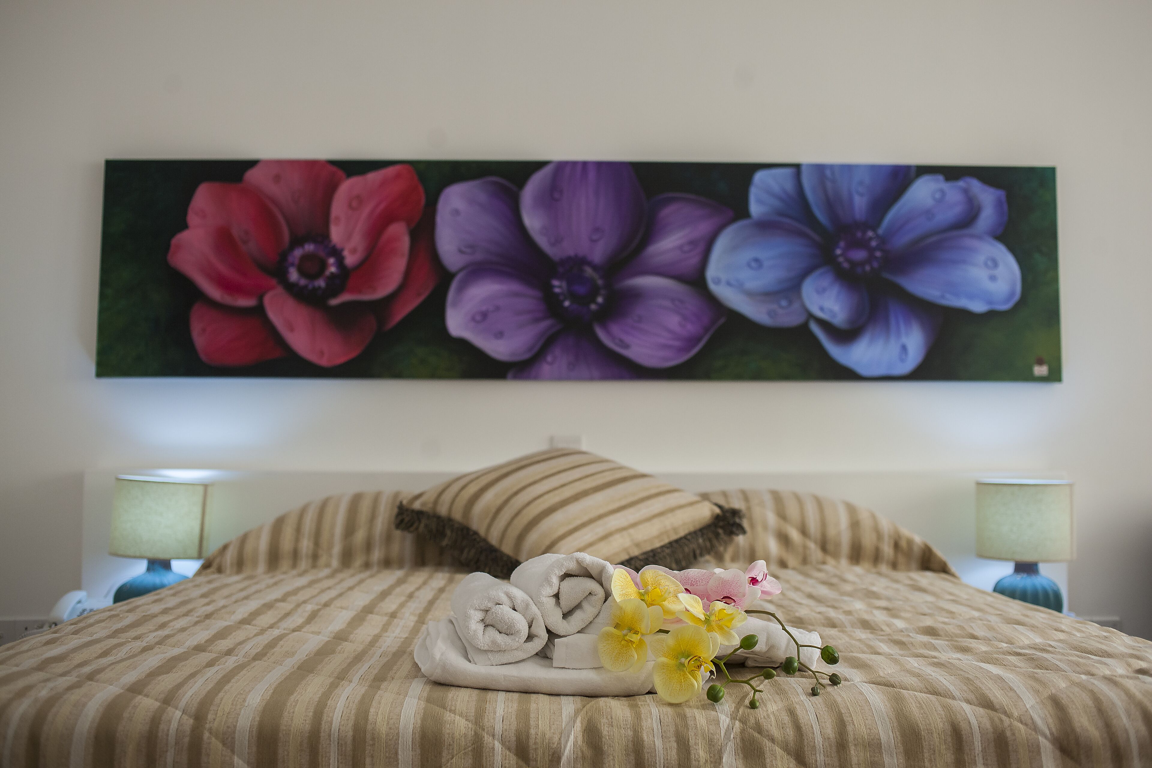 A neatly made bed with striped bedding, a floral painting above, and two bedside lamps.