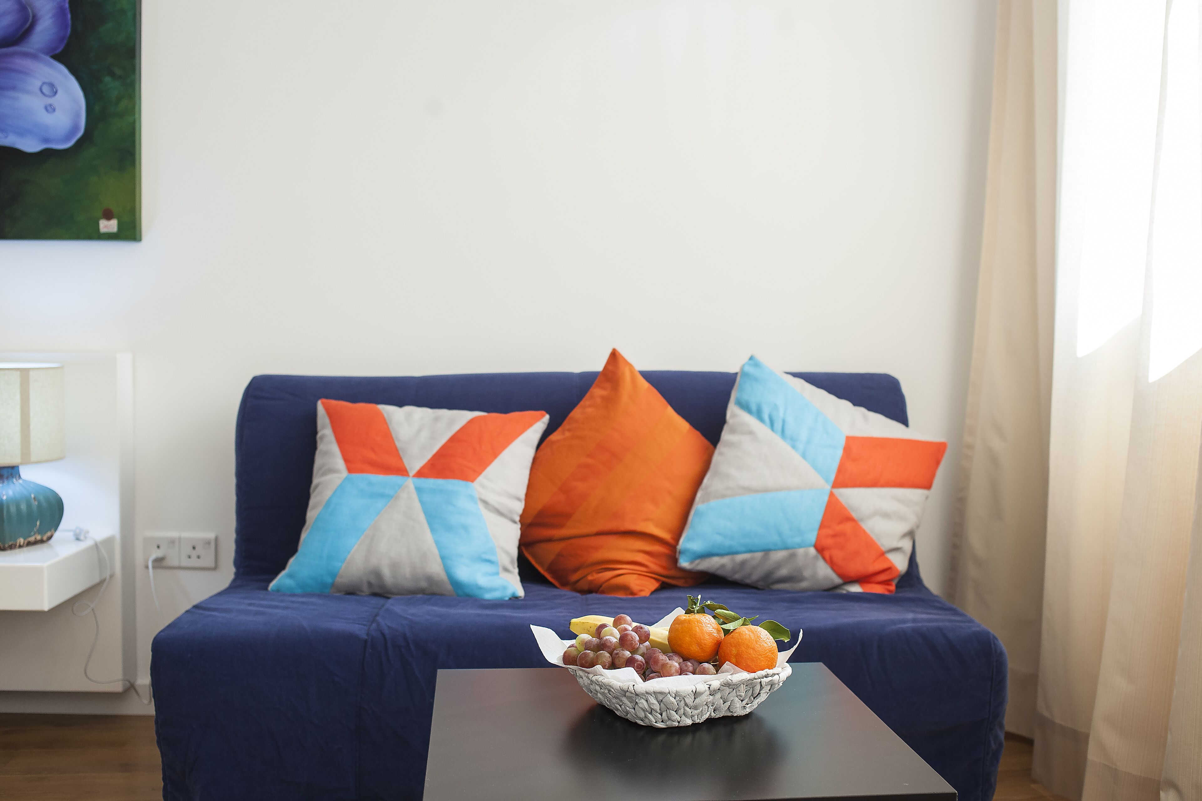 Un canapé bleu orné de coussins à motifs géométriques, un panier de fruits posé sur une table et un tableau accroché au mur.