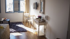 A room with a bed, a desk, a chair, a small table with a bowl of fruit, a potted plant, and a window with curtains. - Operetta House (Larnaca)