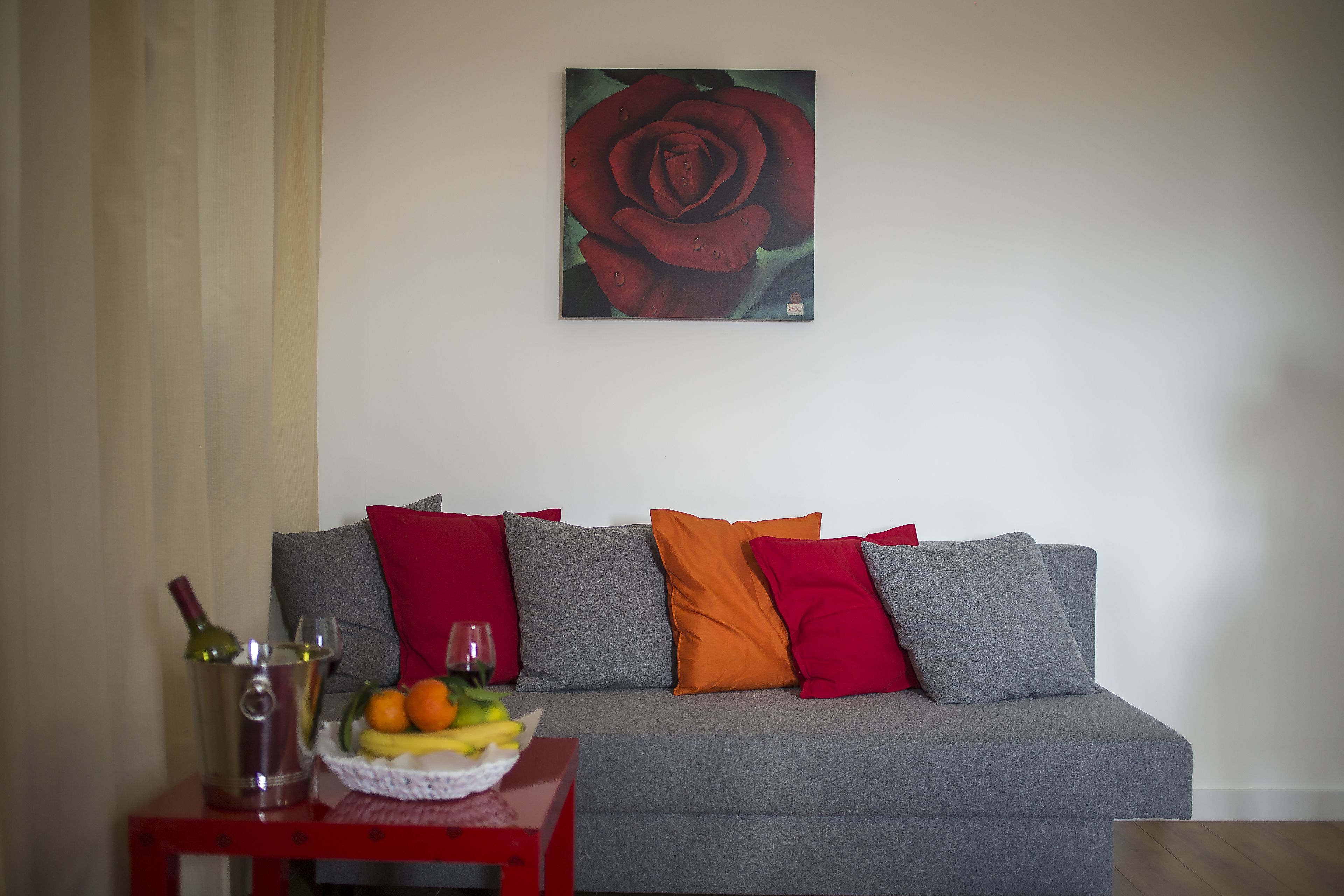 A living room with a grey sofa, red and orange cushions, a small table with a wine bottle, glasses, and a fruit bowl.