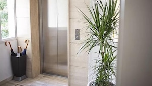 A modern elevator with a glass door, a potted plant beside it, and a trash bin with umbrellas. - Operetta House (Larnaca)