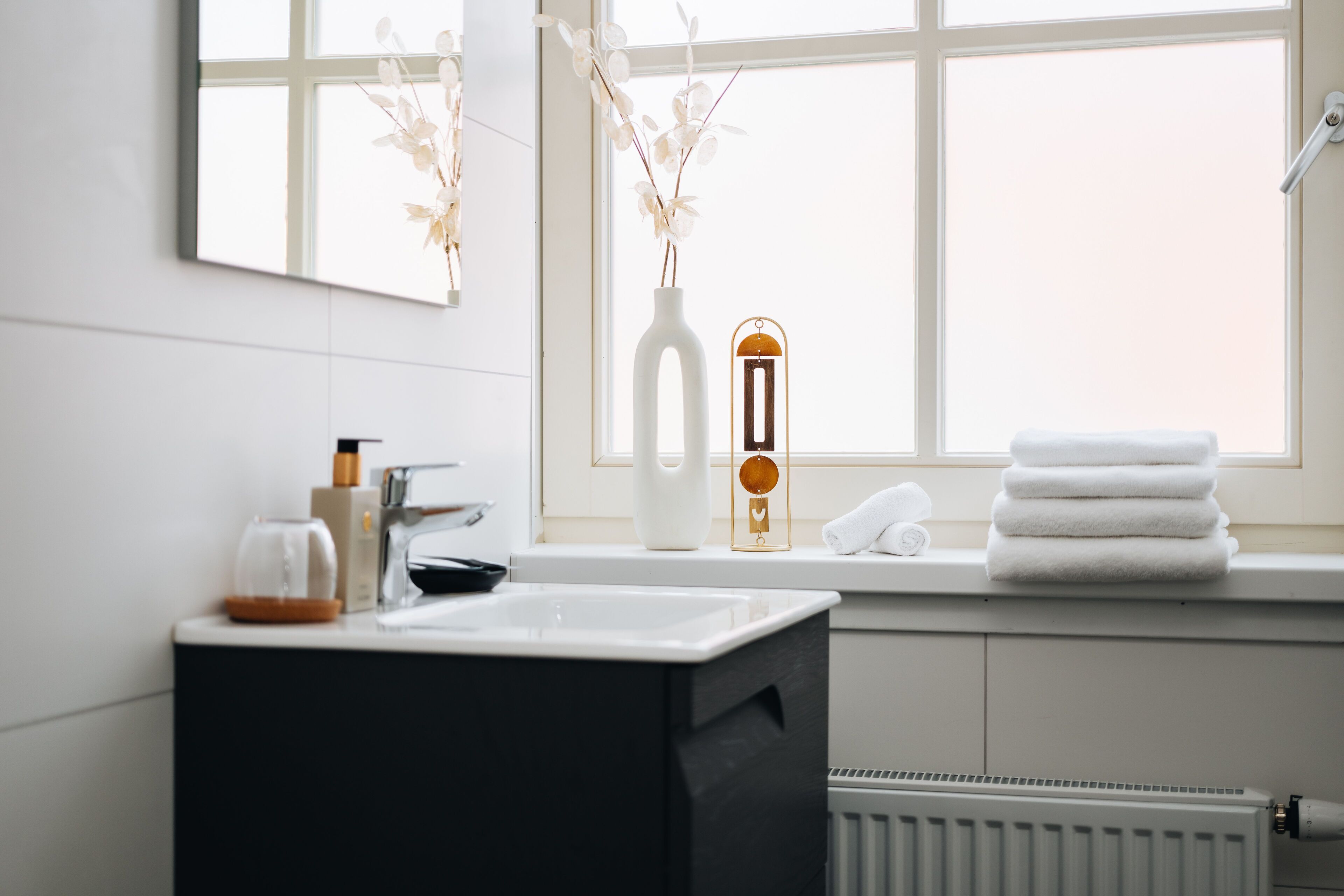 A bathroom with a white sink, a black vanity, a white vase with branches, a thermometer, and towels on the windowsill.