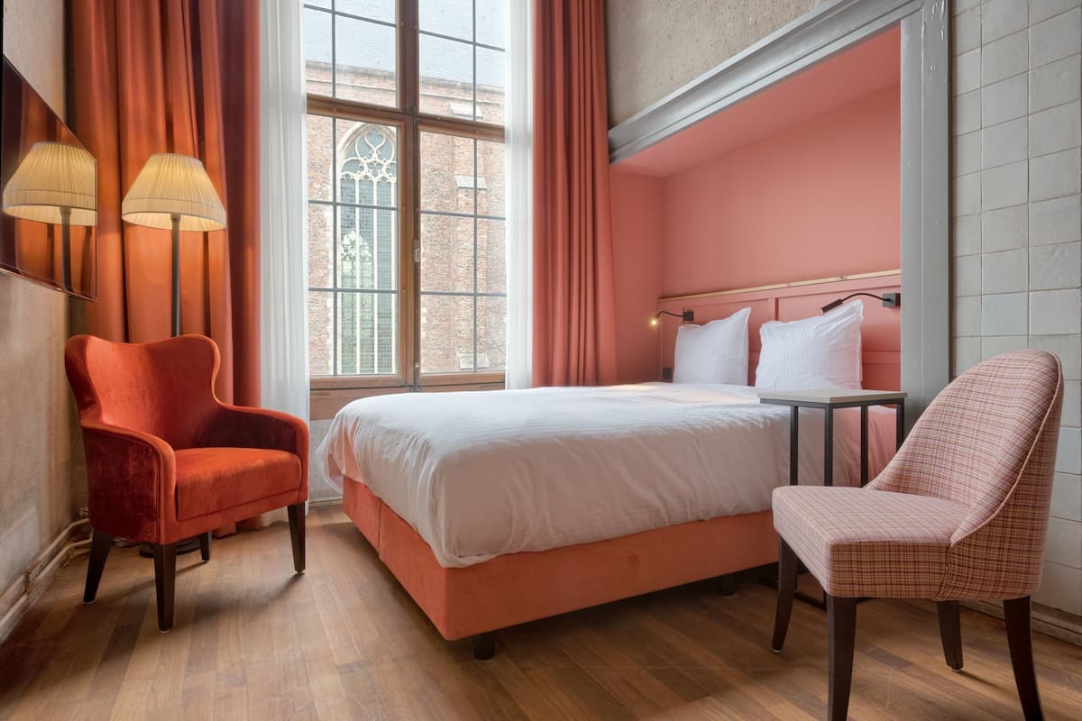 Une chambre d’hôtel avec un grand lit, deux chaises, une table et une fenêtre avec des rideaux.