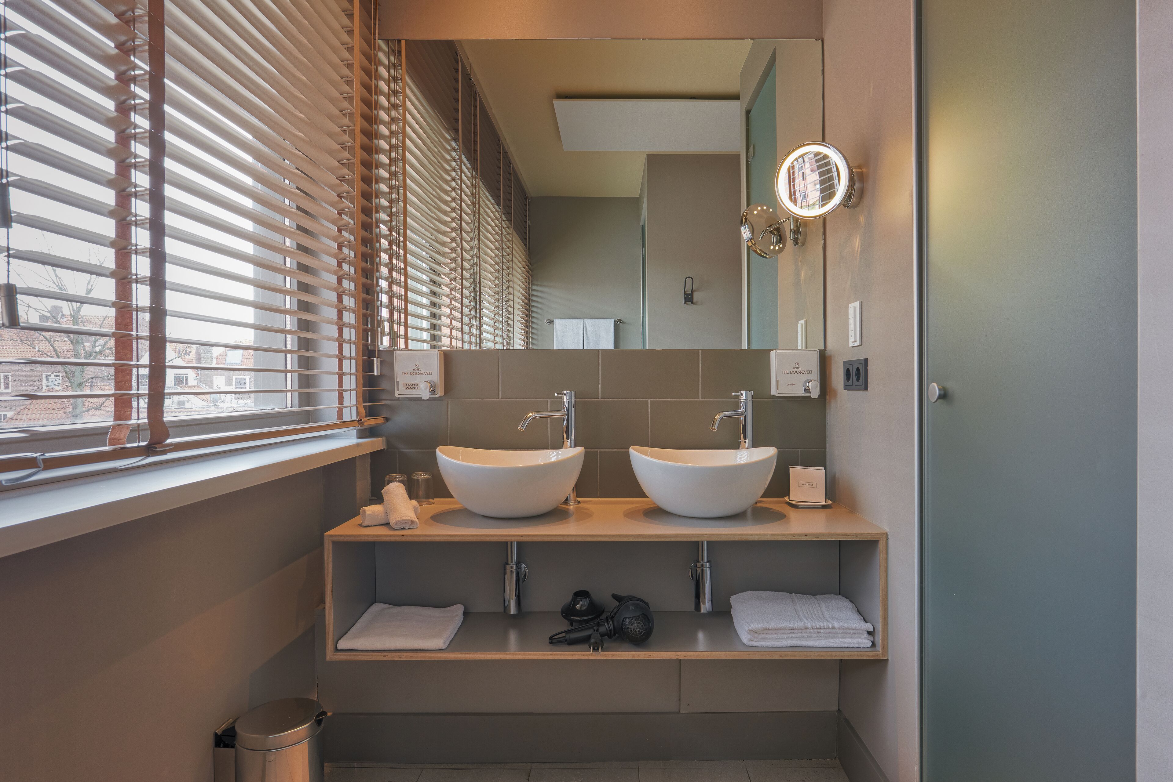 Un baño moderno con dos lavamanos, un espejo y un ventanal con persianas.