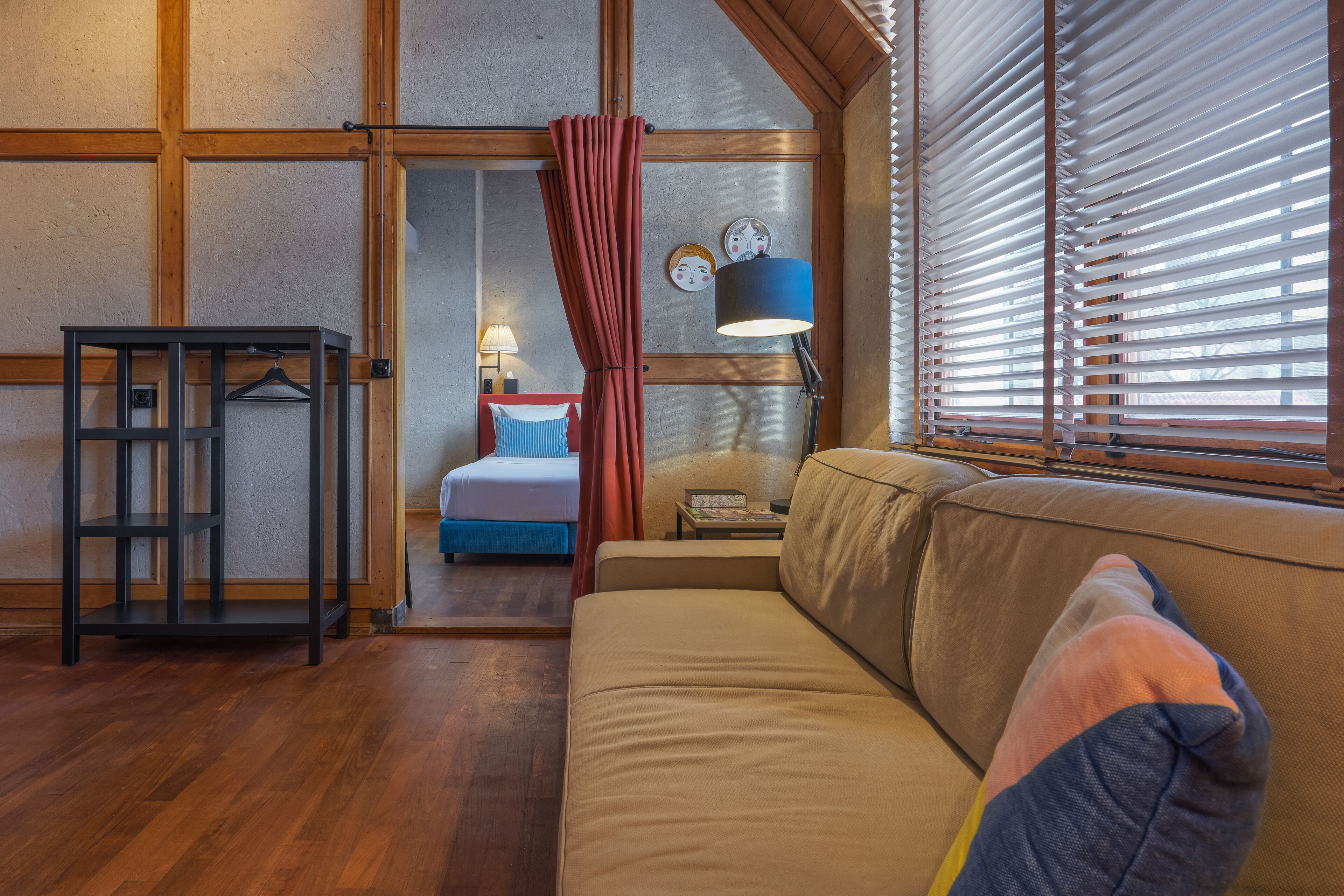 Une pièce chaleureuse avec un plancher en bois, un canapé beige, une petite table, un porte-vêtements suspendu, un lit avec une tête de lit bleue et une fenêtre avec des stores.
