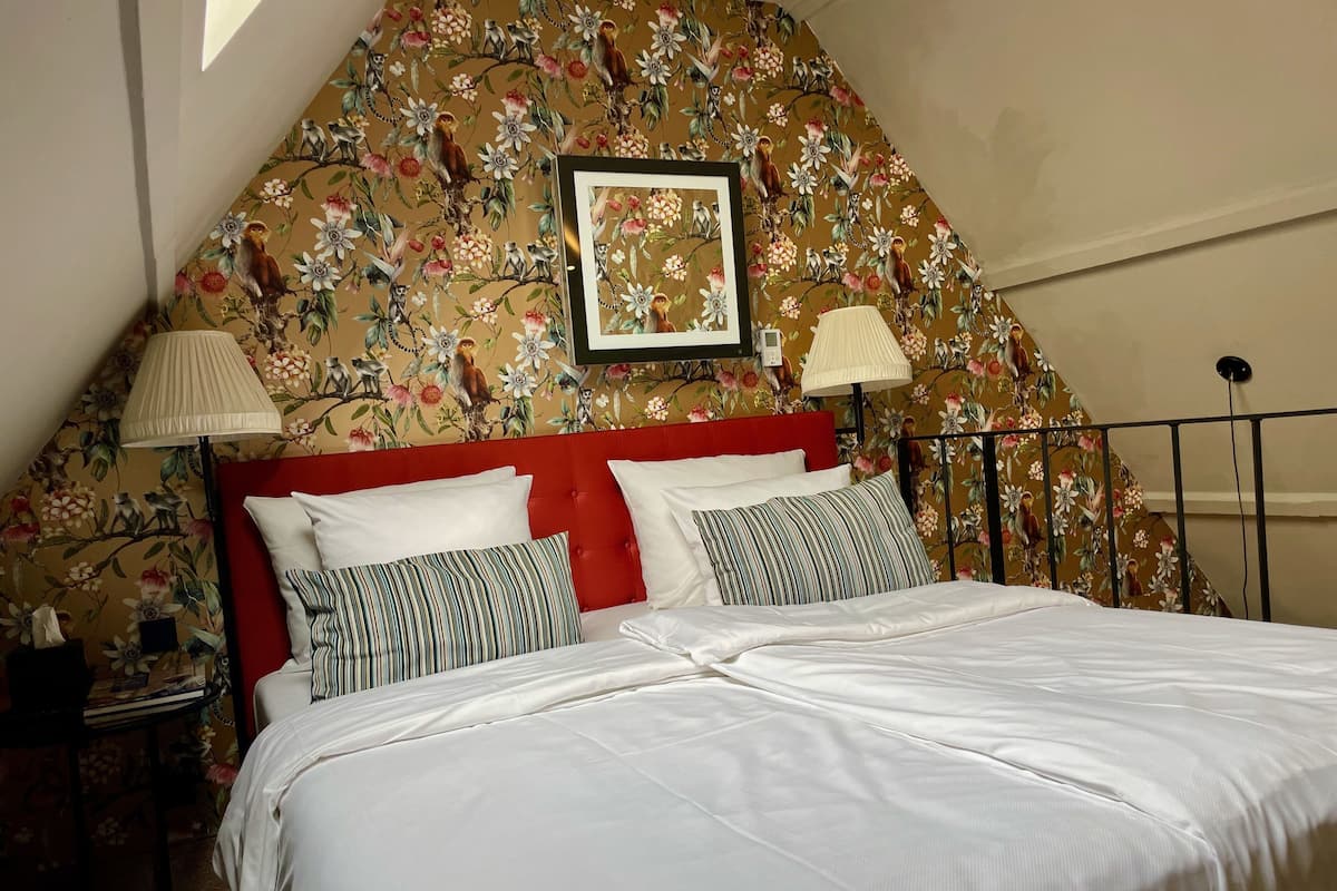 Une chambre à coucher avec une tête de lit rouge, un lit avec une literie blanche, deux lampes et un papier peint à motifs floraux.