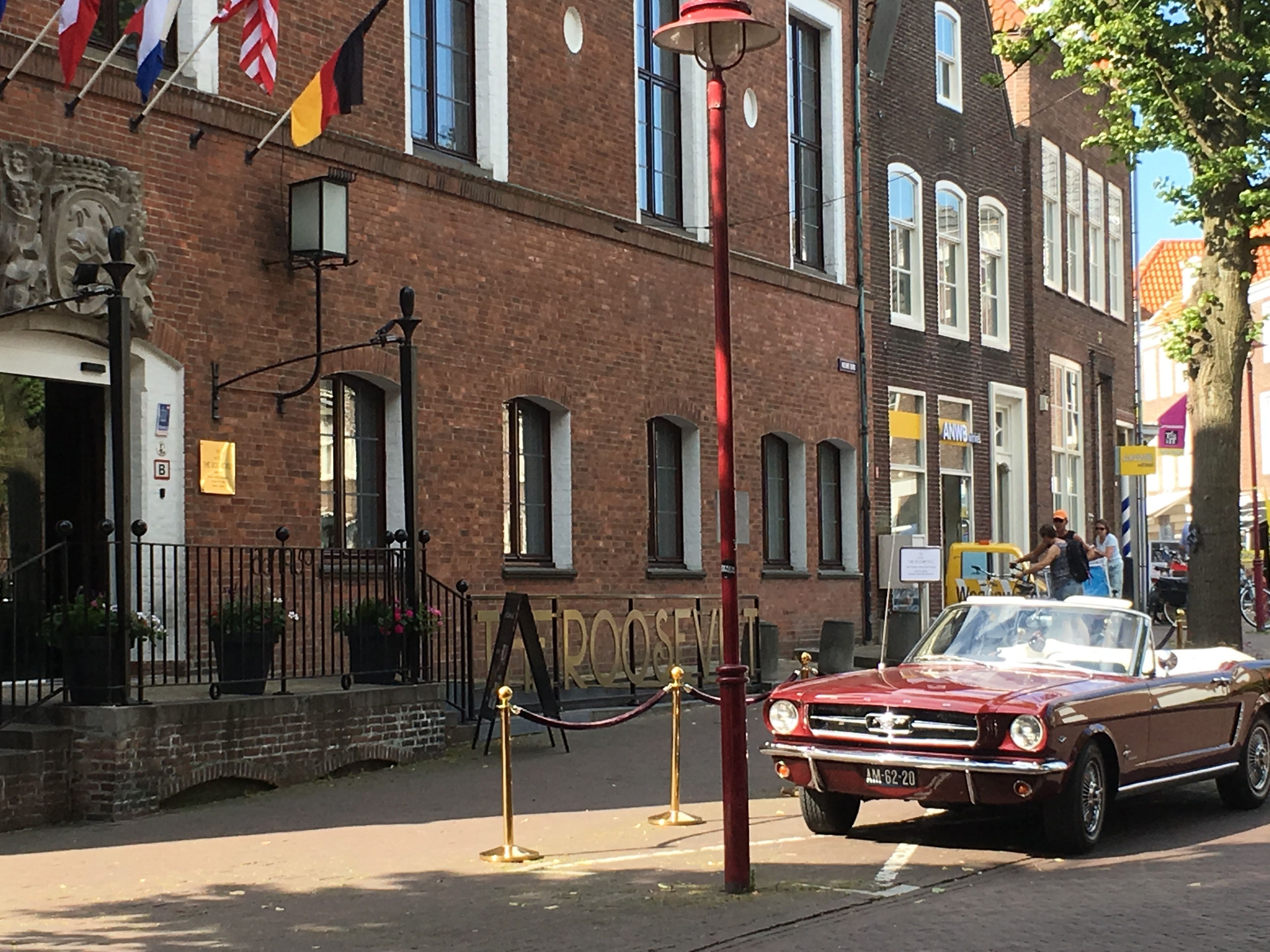 A vintage red convertible parked on a city street with brick buildings and flags in the background.