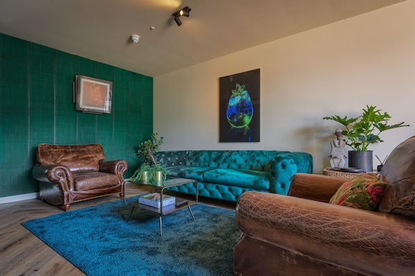 Uma sala de estar com parede verde, sofá verde-água, poltrona de couro marrom, mesa de centro e tapete azul.
